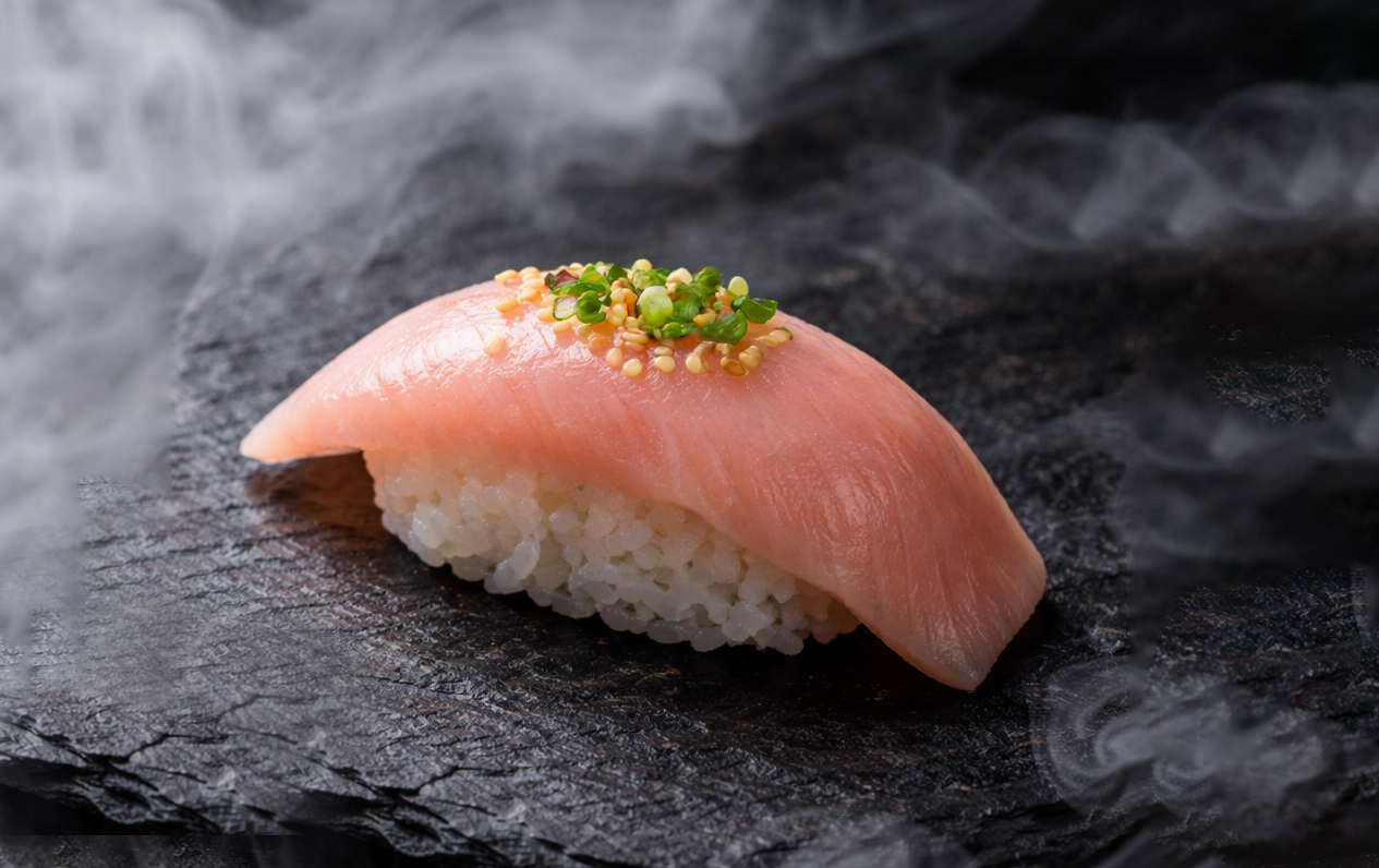 A piece of sushi with a slice of raw fish, topped with chopped green onions and mustard seeds, on a dark, textured slate surface with wisps of smoke.