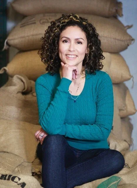 A woman with curly hair sitting on the floor with her arm resting on her knee, smiling, in front of stacked burlap sacks.