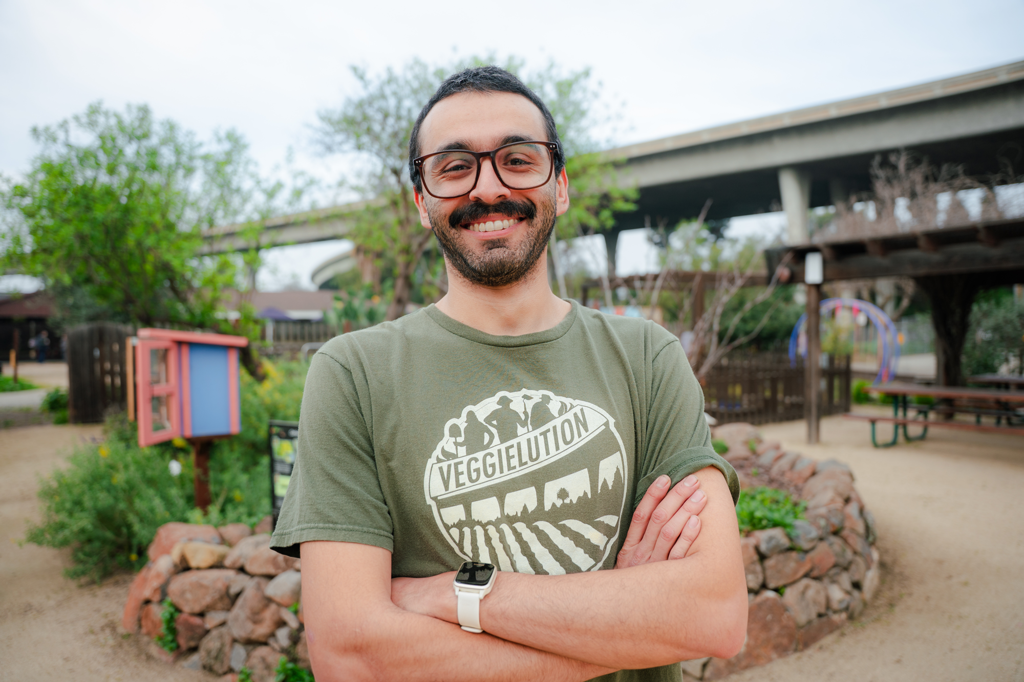 A smiling man with glasses and a beard wearing a green VeggieLution T-shirt standing outdoors with arms crossed at a park.