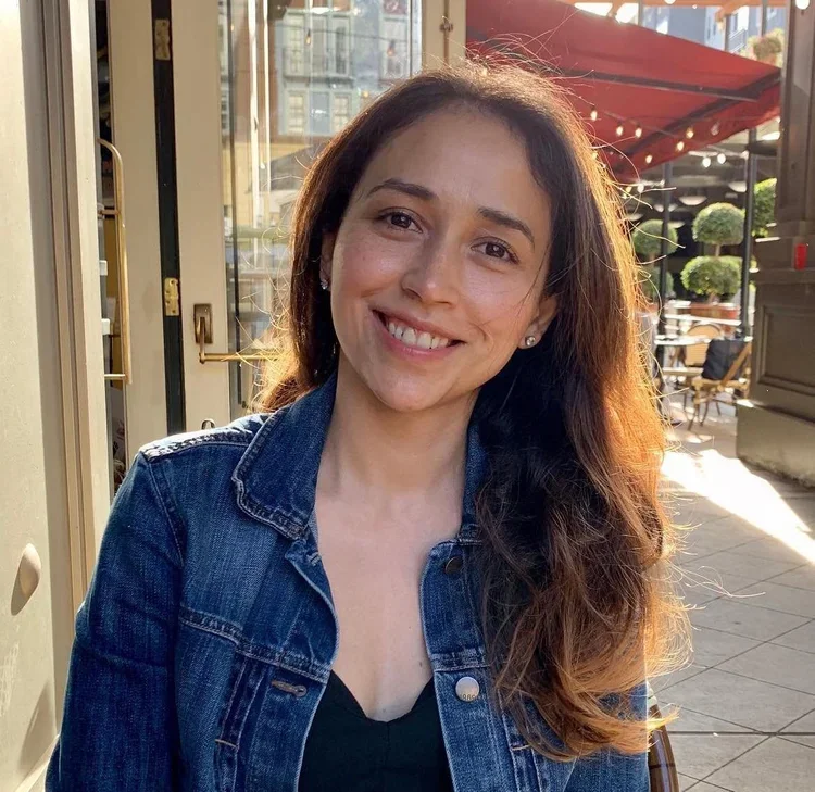 Smiling young woman with long wavy hair, wearing a denim jacket and black top, sitting outdoors at a cafe during sunset.