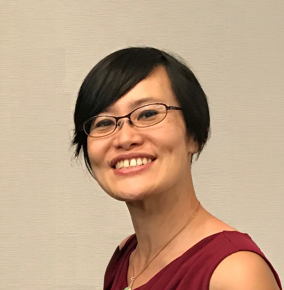 A smiling woman with short black hair, glasses, and light skin, wearing a sleeveless maroon top and a gold necklace.