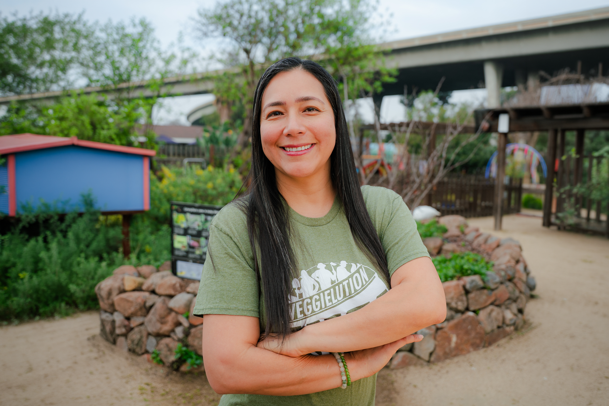 Woman with long black hair smiling, standing outdoors in a garden with trees, a wooden structure, and a bridge in the background.