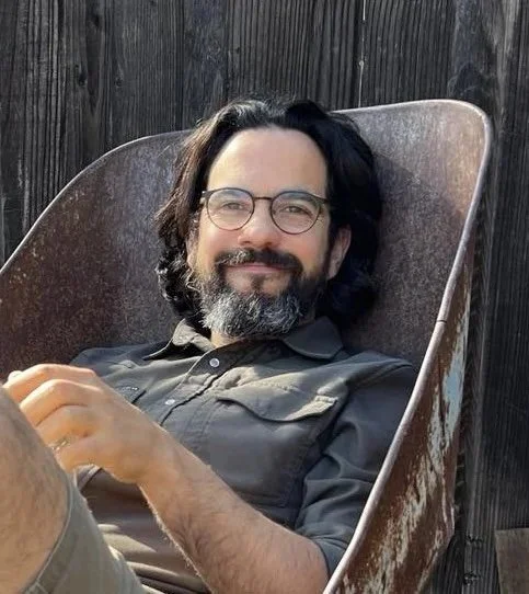 A man with glasses and a beard reclining in a metal outdoor chair, smiling, with a wooden fence in the background.