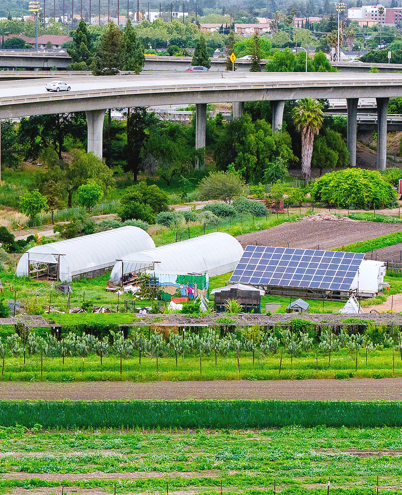 A scenic view of a multi-layered highway overpass surrounded by green trees, cultivated farmland, and a distant city with mountains in the background under a cloudy sky.
