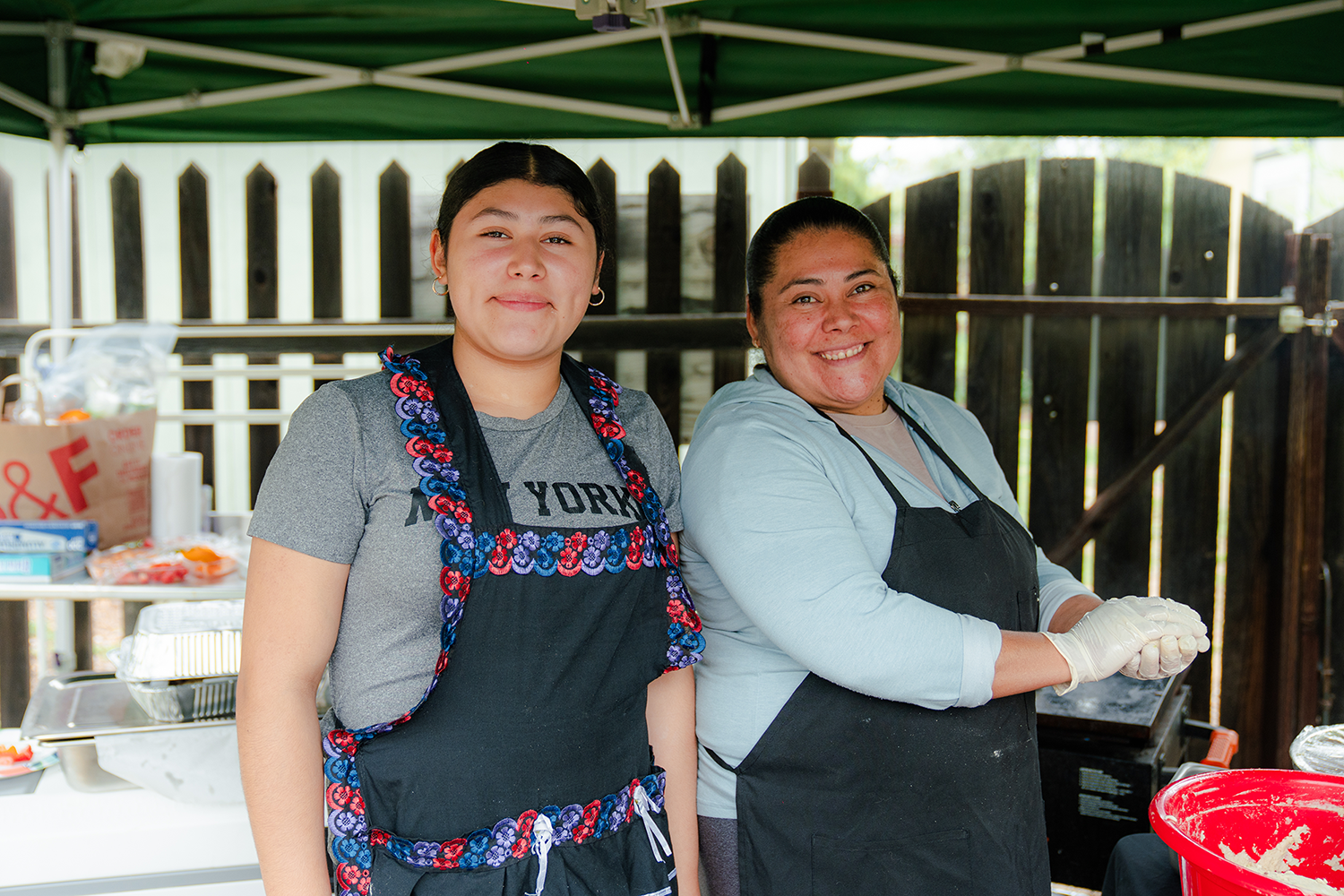 Two women standing under a green canopy at an outdoor cooking or food event, smiling at the camera, with tables and food items in the background.