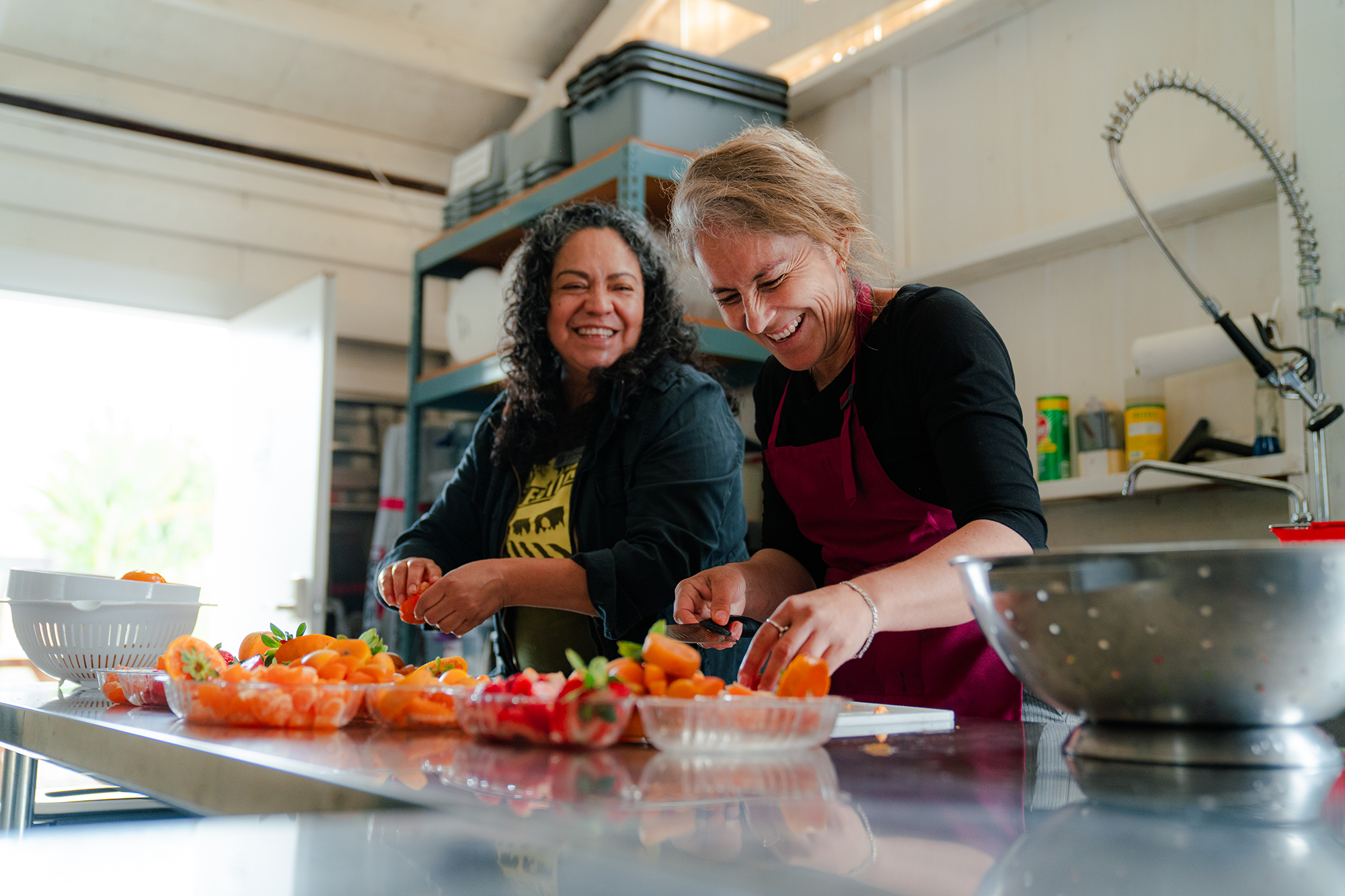 Two women smiling and preparing strawberries in a kitchen with fresh strawberries on the table.