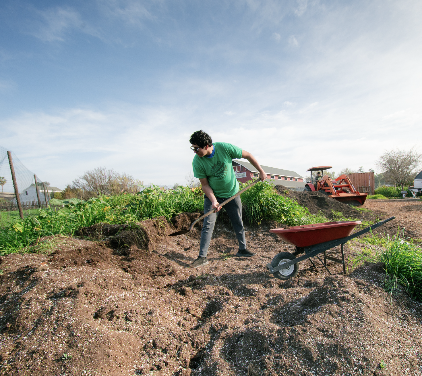 Person working in a farm garden, shoveling soil with a wheelbarrow nearby, under a partly cloudy sky.