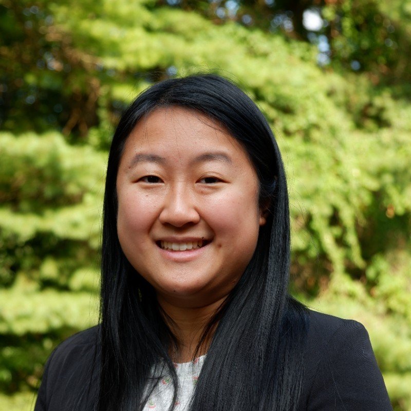 A woman with long black hair smiling outdoors, with green trees in the background.