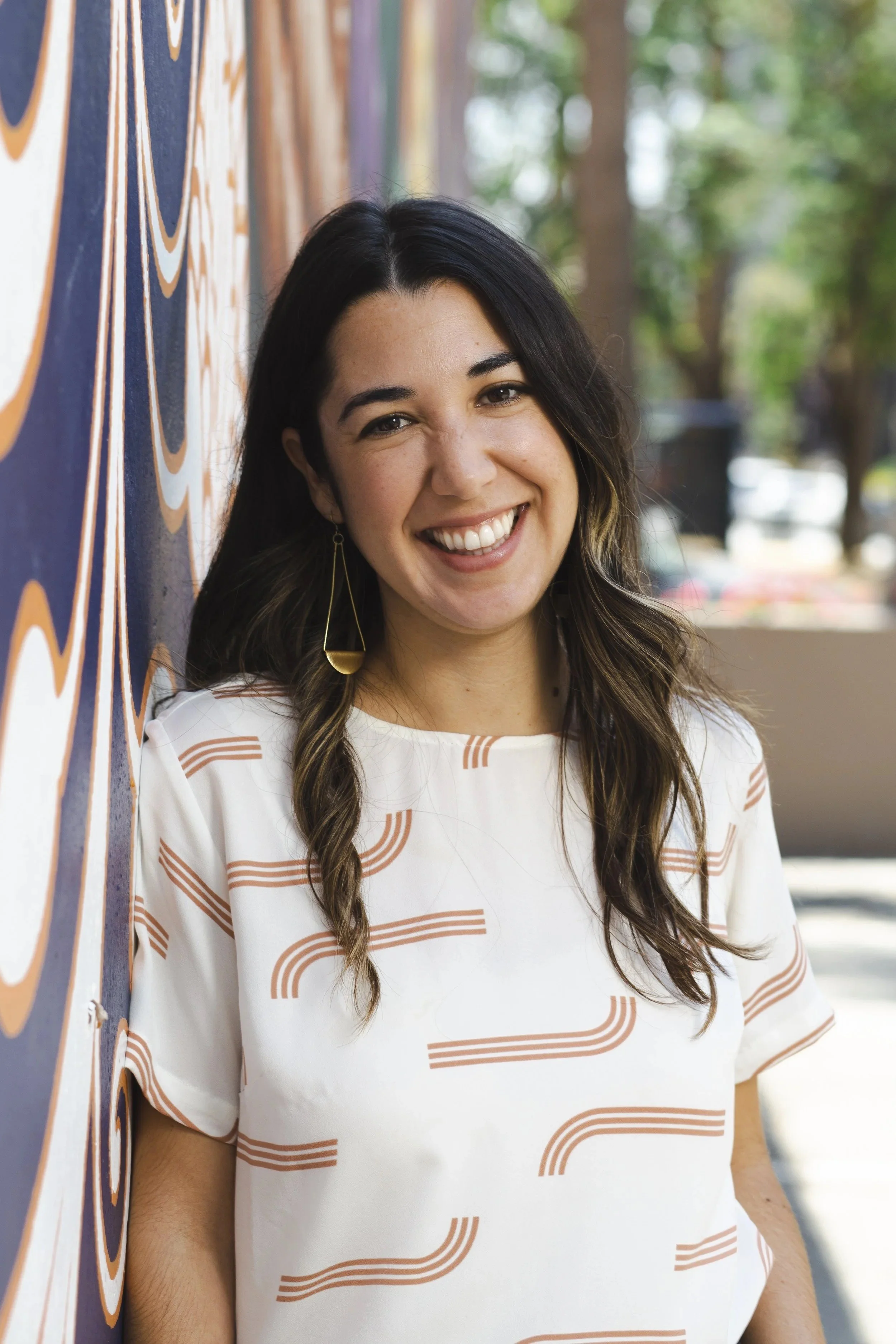 A woman with dark hair and a white shirt with orange curved lines, smiling and standing outdoors next to a colorful mural and trees in the background.