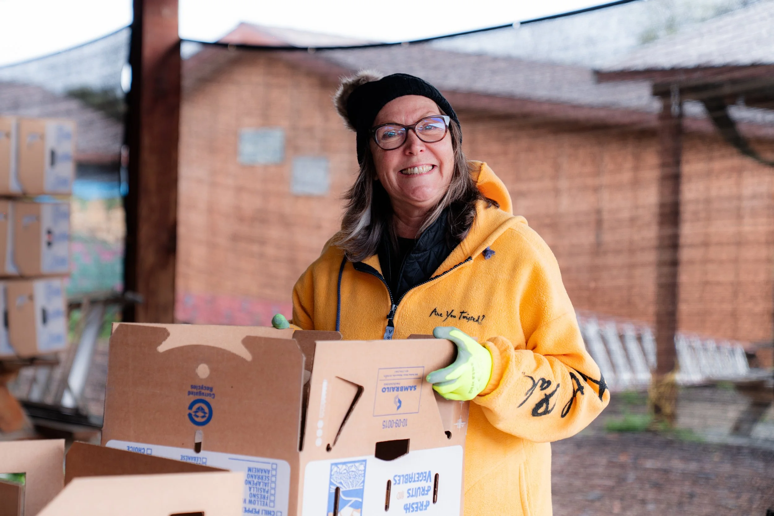 A woman wearing glasses, a black beanie, and a yellow hoodie is holding a cardboard box outdoors. She is smiling and wearing yellow gloves. The background includes a brick building and stacked boxes.