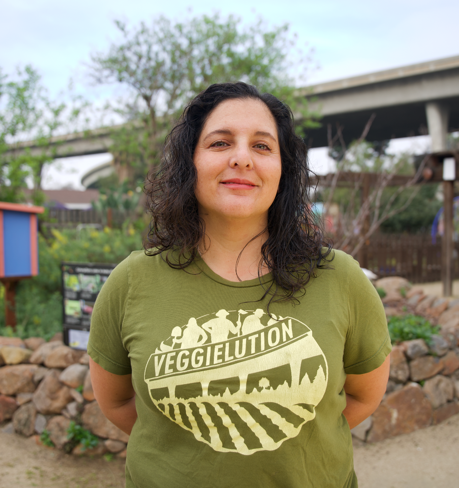 A woman with curly dark hair and a light skin tone, wearing a green t-shirt with a VeggieLution logo, stands outdoors in a park with trees, rocks, and a bridge in the background.