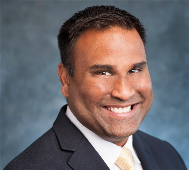 Professional headshot of a smiling man wearing a dark suit, white shirt, and light-colored tie, with a neutral background.