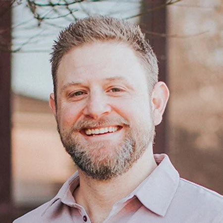 A smiling man with short brown hair and a beard, wearing a light purple collared shirt, outdoors.
