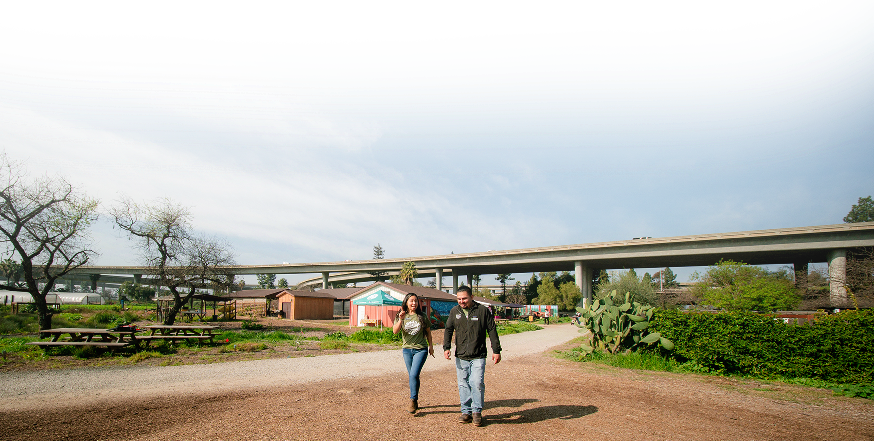 Two people walking together on a dirt path in a park with trees, small structures, and a cactus, with a large highway overpass in the background under a partly cloudy sky, featuring some digital artifacts or distortions in the sky.