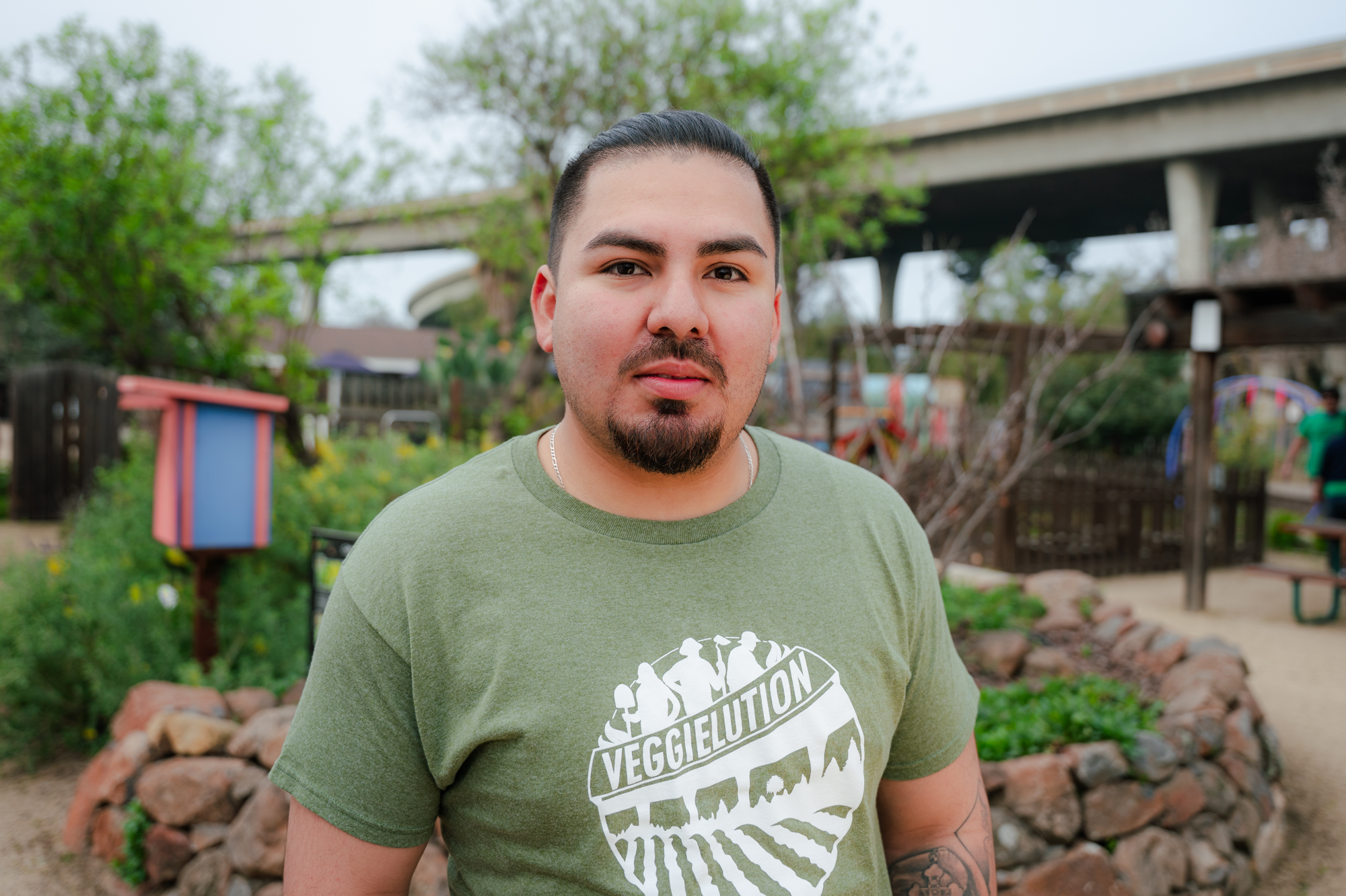 A man with dark hair, a goatee, wearing a green T-shirt with white text, stands outdoors in front of a park or garden with trees, a small structure, and children in the background.