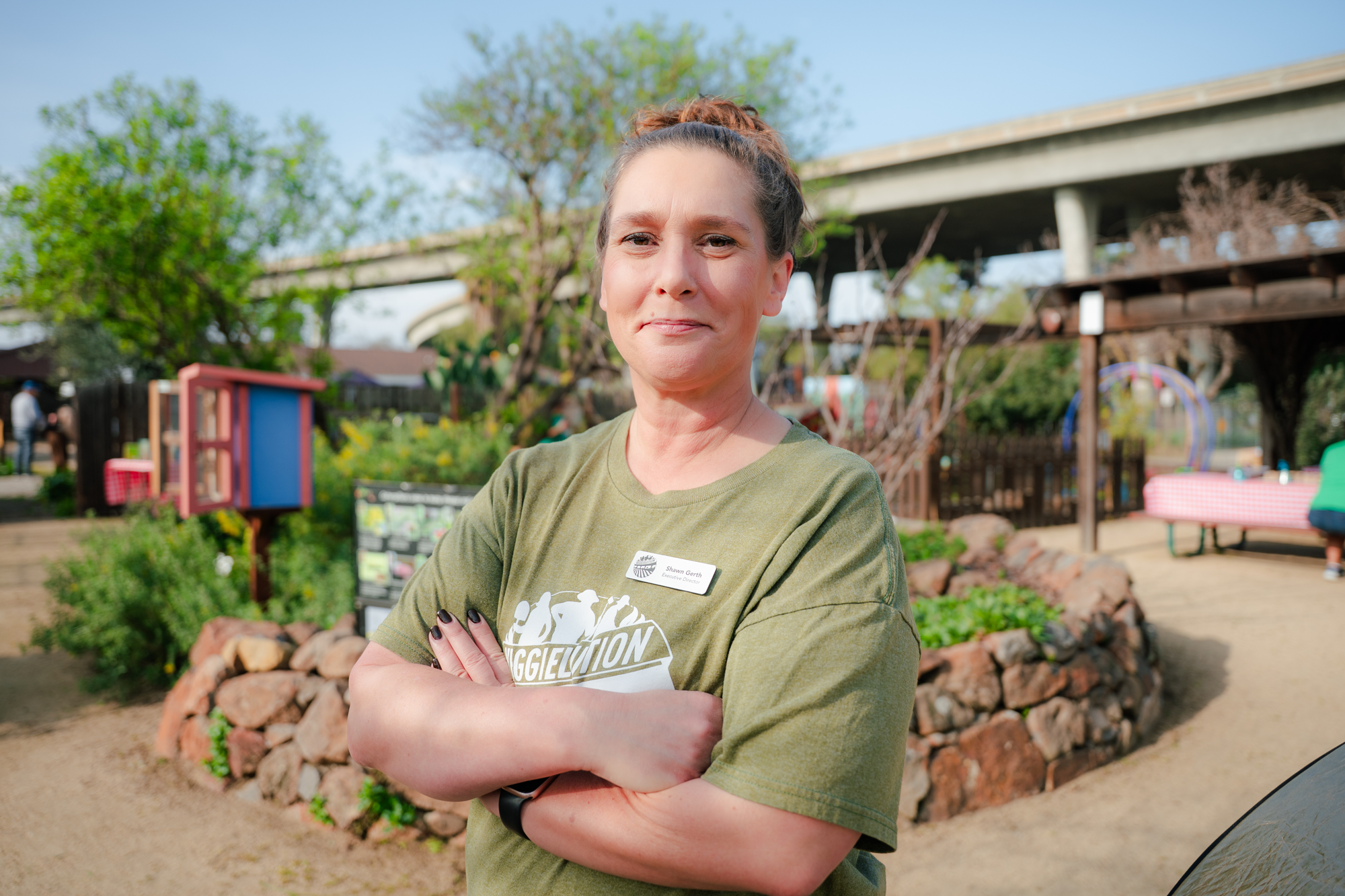 A woman with brown hair in a bun, wearing a green T-shirt and a name tag, standing with arms crossed in an outdoor park setting during daytime.