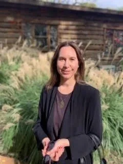 A woman standing outdoors in front of a rustic wooden building with tall grass and plants around her.