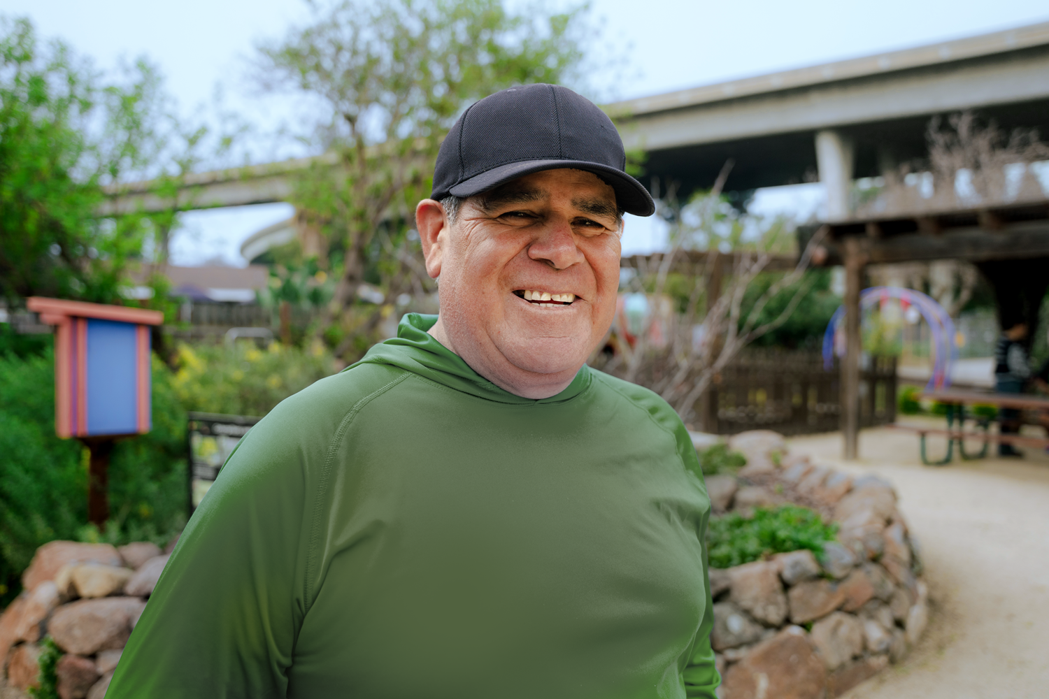 A smiling man in a green shirt and black cap standing outdoors in a park or playground with trees, a bridge, and playground equipment in the background.