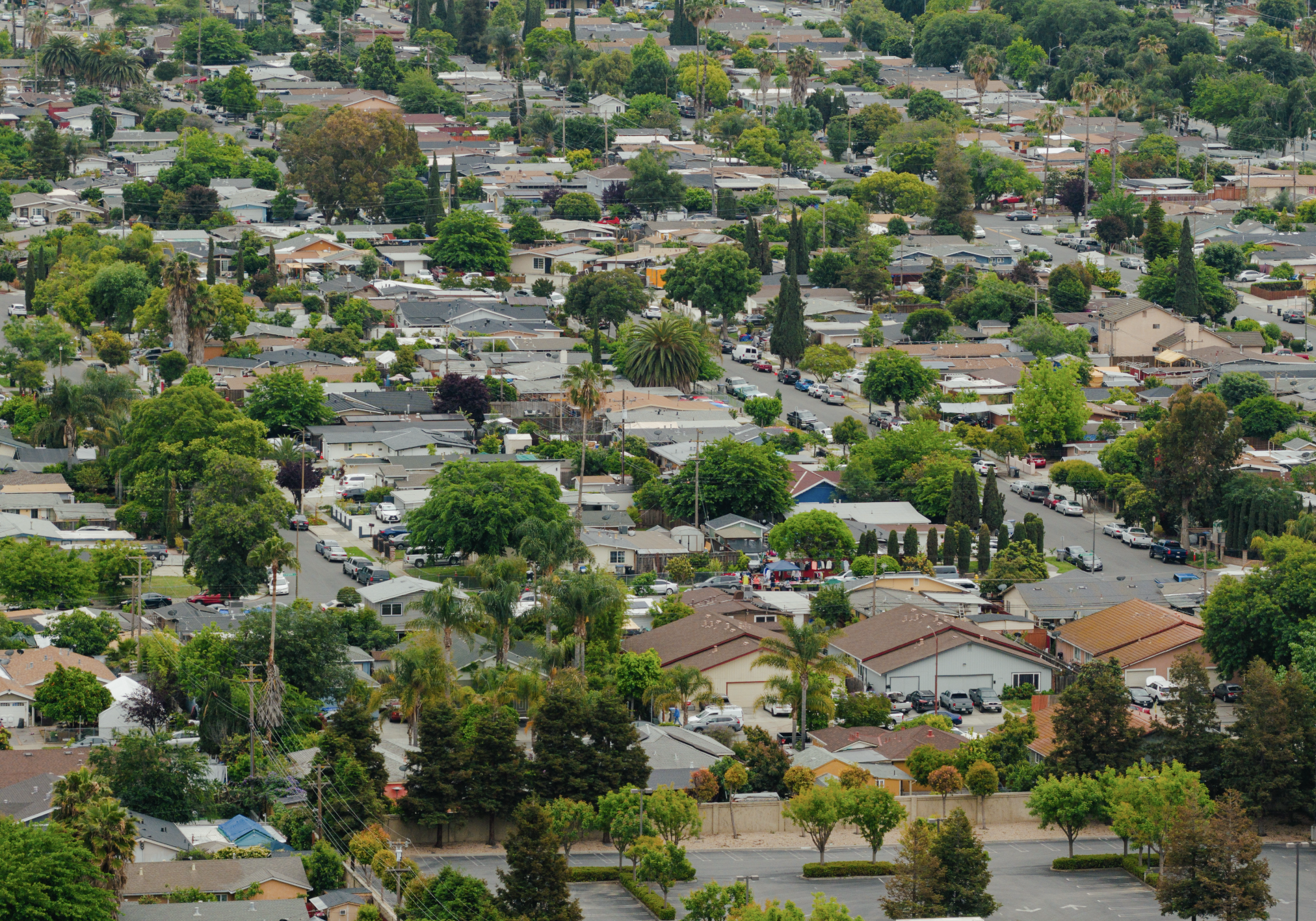 Aerial view of a suburban neighborhood with houses, trees, and streets, and a large parking lot in the lower part of the image.