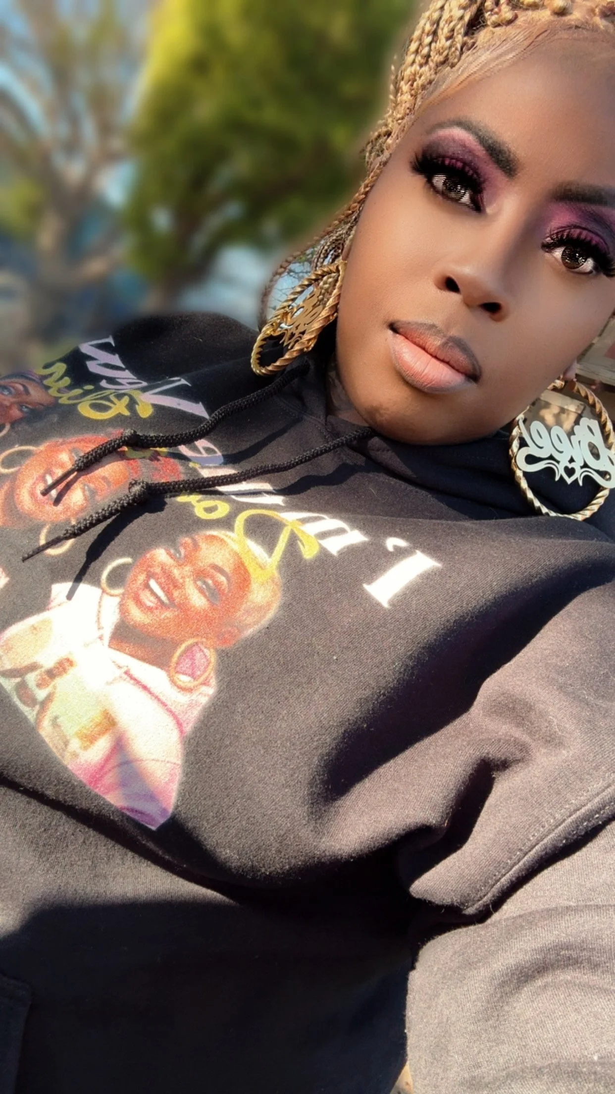 A woman with braided blonde hair, wearing large gold hoop earrings and vibrant makeup, taking a selfie outdoors on a sunny day.