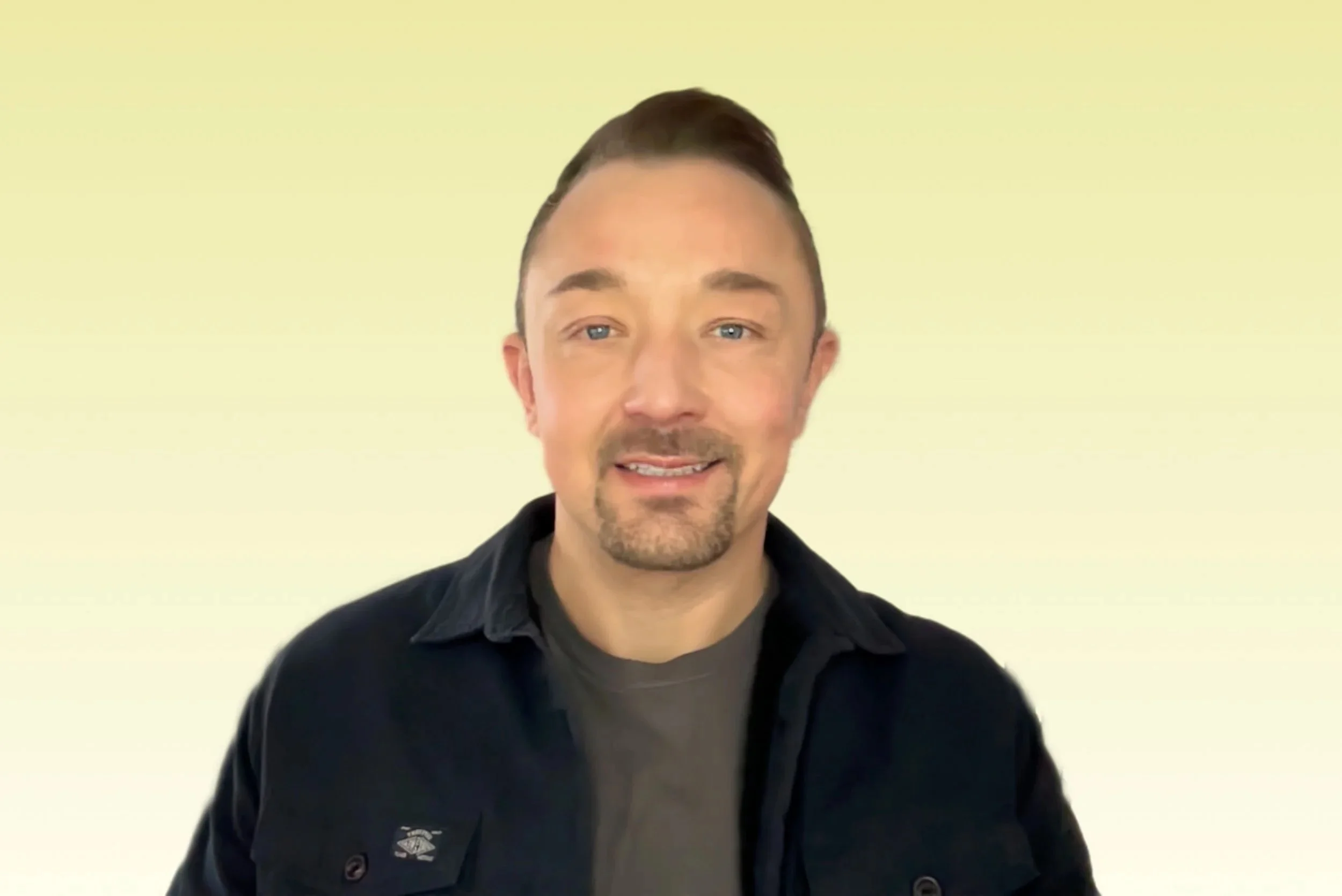 A man with short brown hair, a goatee, and blue eyes, smiling, wearing a dark shirt over a gray t-shirt, standing against a light yellow background.