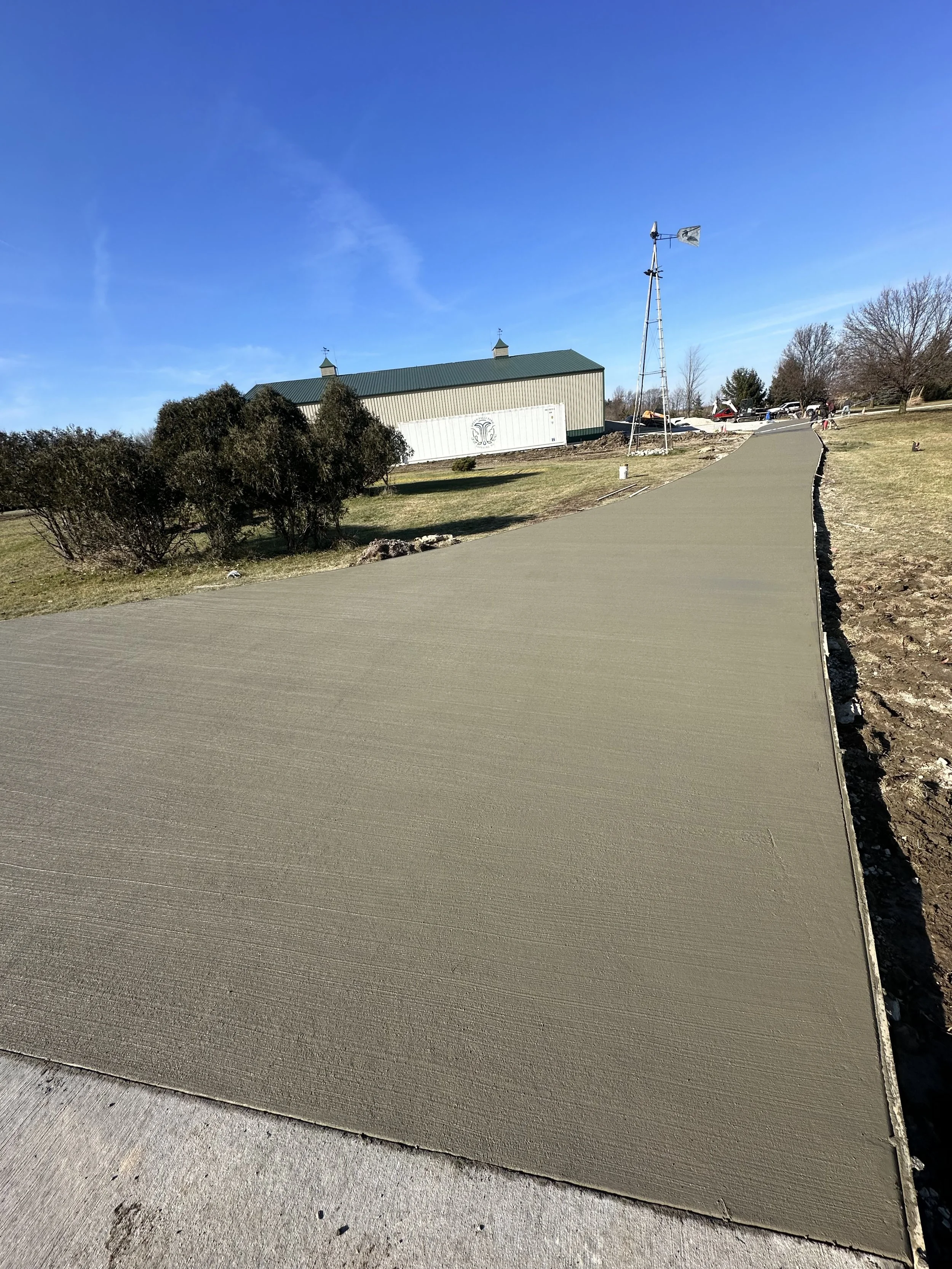 A freshly poured concrete sidewalk on a clear, sunny day, leading towards a building with a green roof and a windmill in the background.