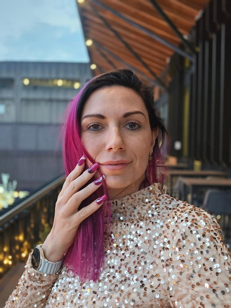 A woman with pink hair wearing a sparkly, sequin-covered top, posing with her hand near her face, in an indoor setting with a wooden ceiling and windows in the background.