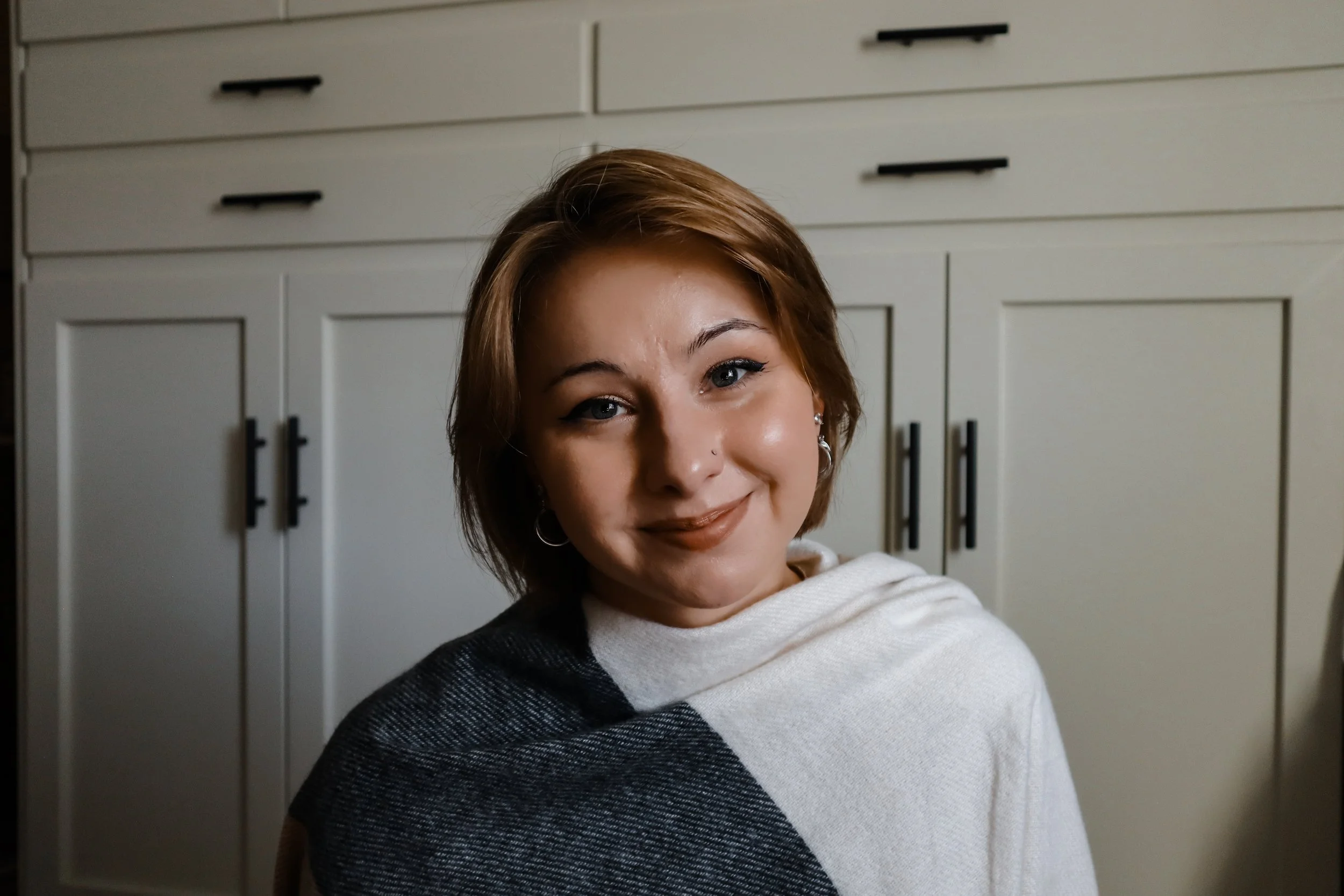 A woman with short brown hair, light skin, and earrings smiling at the camera, sitting in front of white kitchen cabinets.