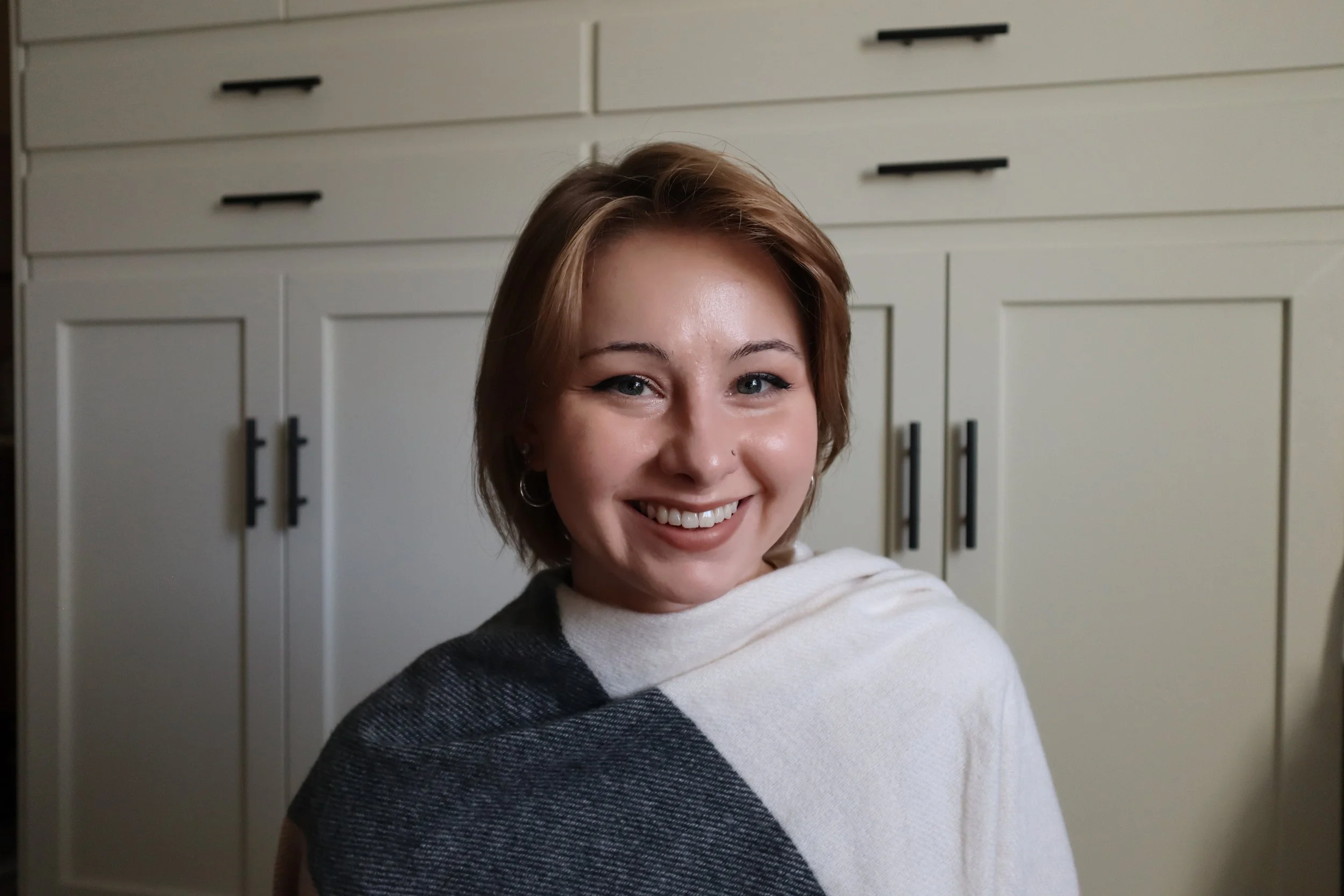 A smiling woman with light brown hair and hoop earrings, wearing a black and white top, in front of white kitchen cabinets.