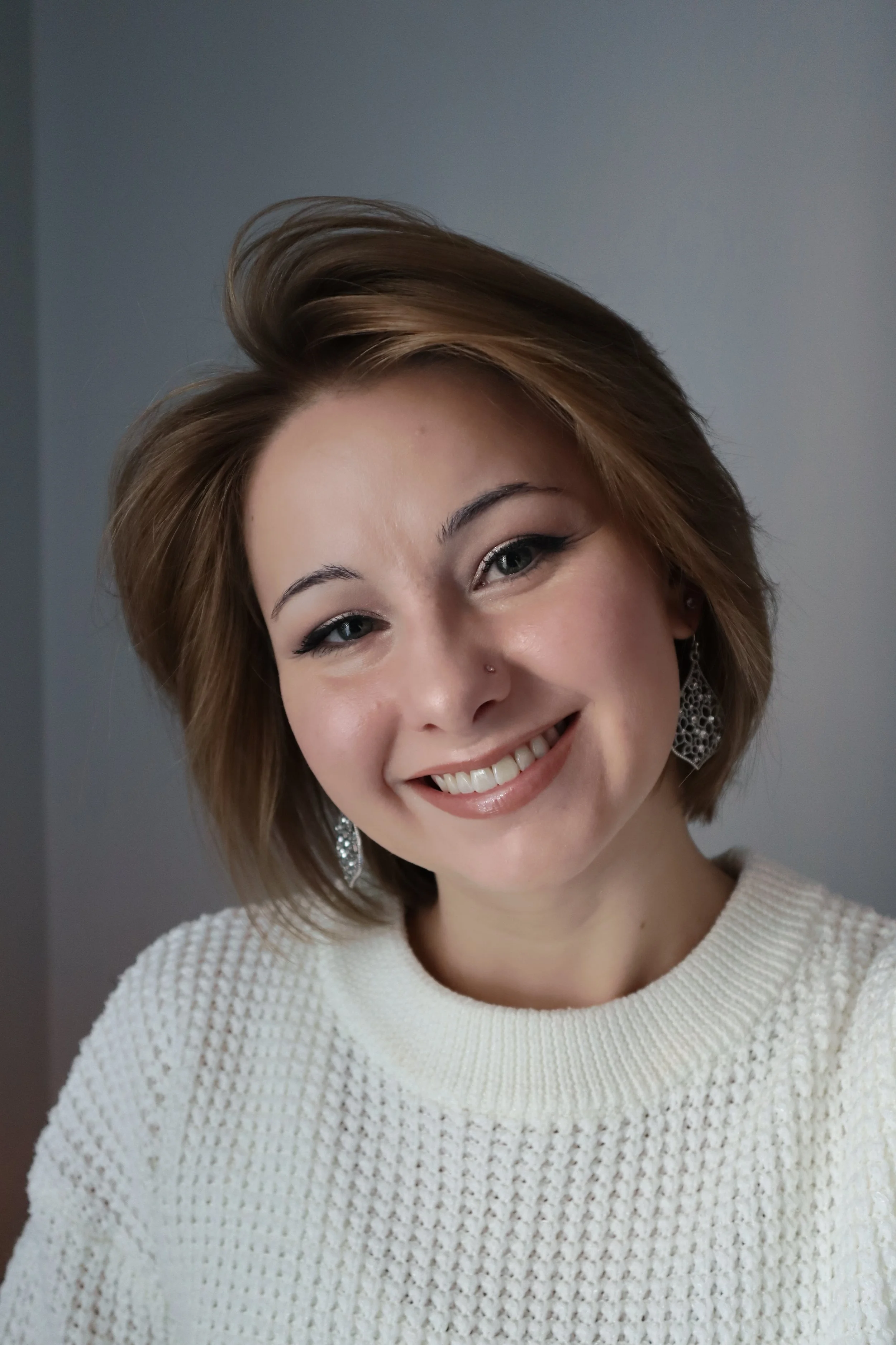 Close-up of a woman with short, light brown hair smiling and looking at the camera, wearing earrings and a white textured sweater.
