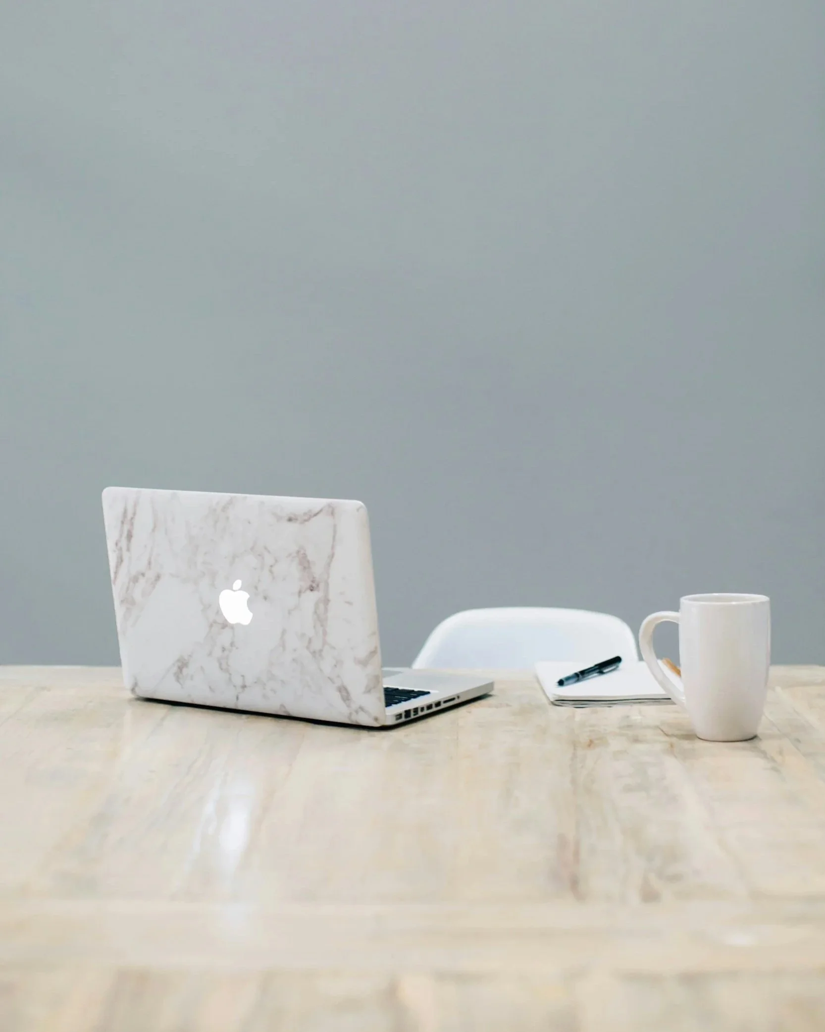 A wooden table with a marble laptop, a white chair, a notepad with a pen, and a coffee mug in front of a plain grey wall.