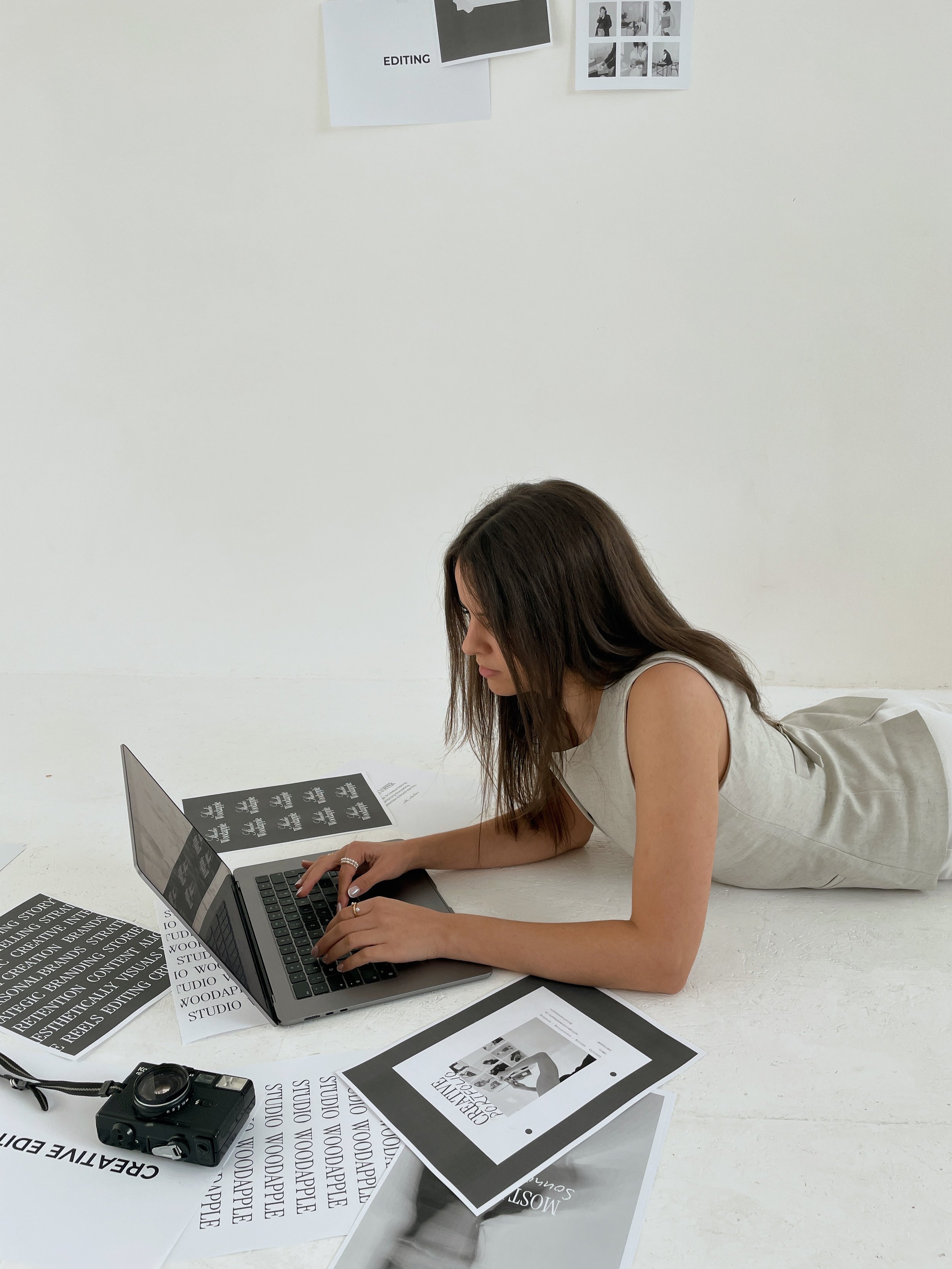 Junge Frau arbeitet auf dem Boden mit Laptop, umgeben von Fotografien, gedruckten Texten und einem Kamera in einem Büro oder Studio.
