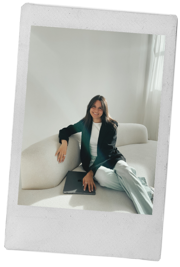 Eine junge Frau sitzt lächelnd auf einer weißen Couch in einem hellen Raum mit großen Fenstern, trägt eine schwarze Jacke und weiße Hose, hat ein Laptop auf dem Schoß.