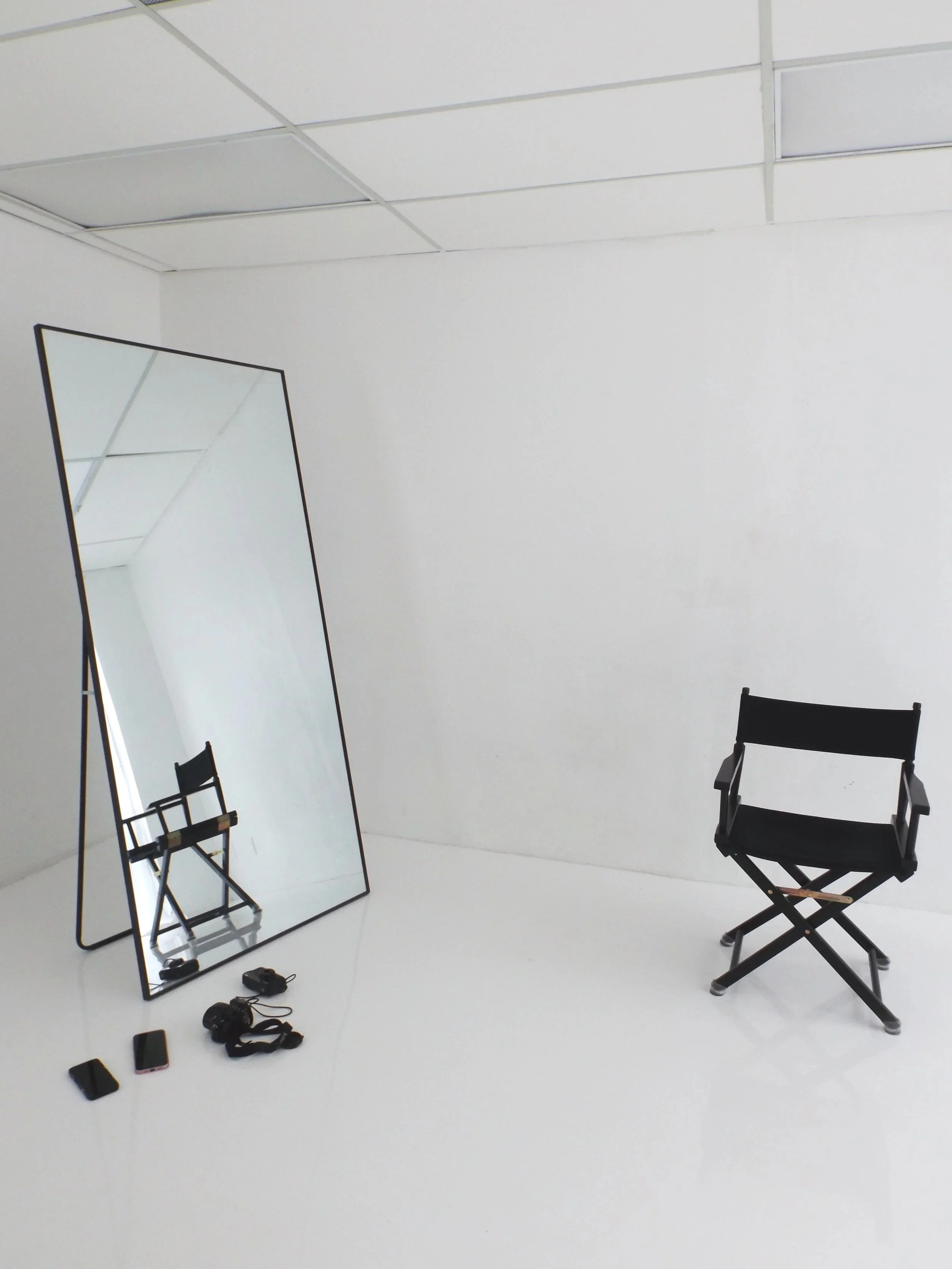 Ein leeres Fotostudio mit einem großen, rechteckigen Spiegel auf dem Boden, einem schwarzen Regiestuhl, und mehreren kleinen Gegenständen am Boden, darunter zwei Mobiltelefone, ein Aschenbecher, eine Kamera und ein Kabel.
