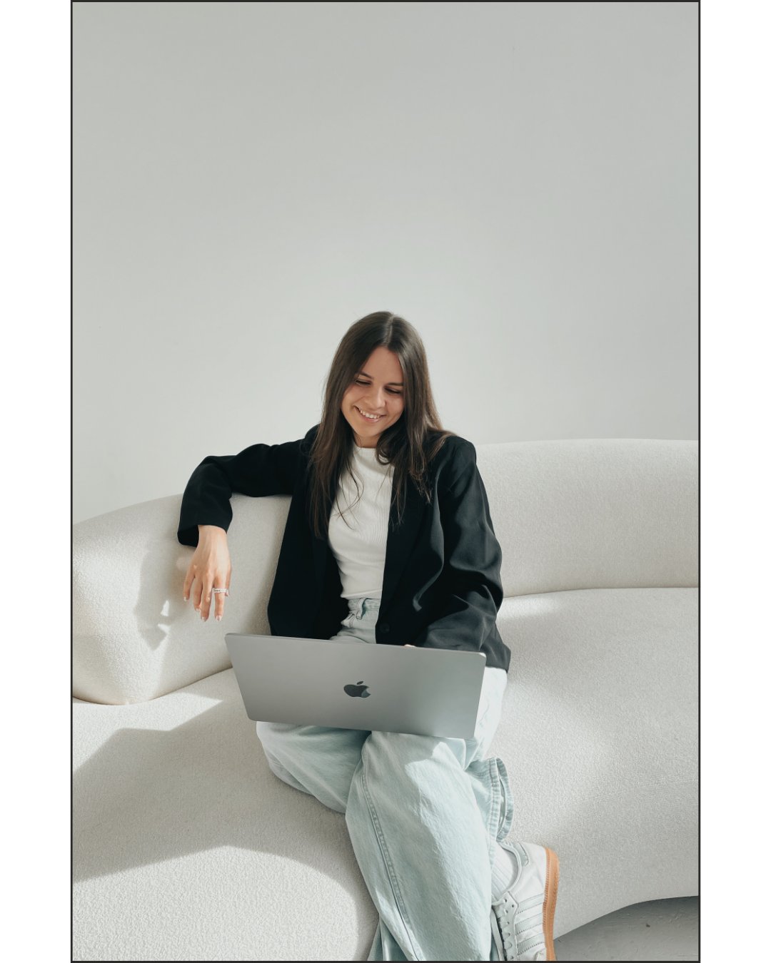 Junge Frau sitzt lachend auf einem weißen Sofa und arbeitet an einem Laptop.