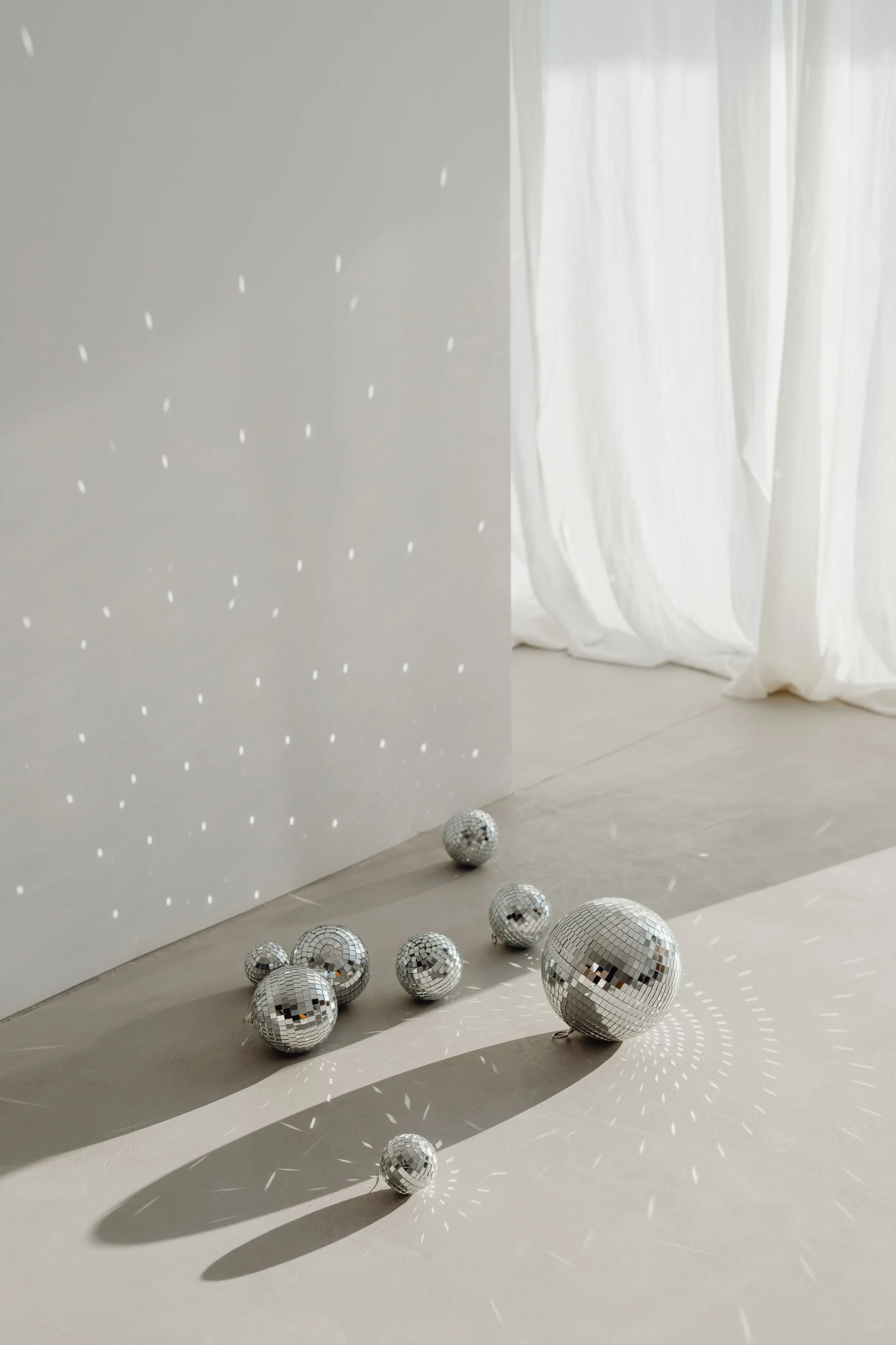 Mehrere kleine und große Disco-Kugeln auf einem Boden, die Lichtreflexe und Muster an die Wand und den Boden werfen, im Hintergrund weiße Vorhänge.