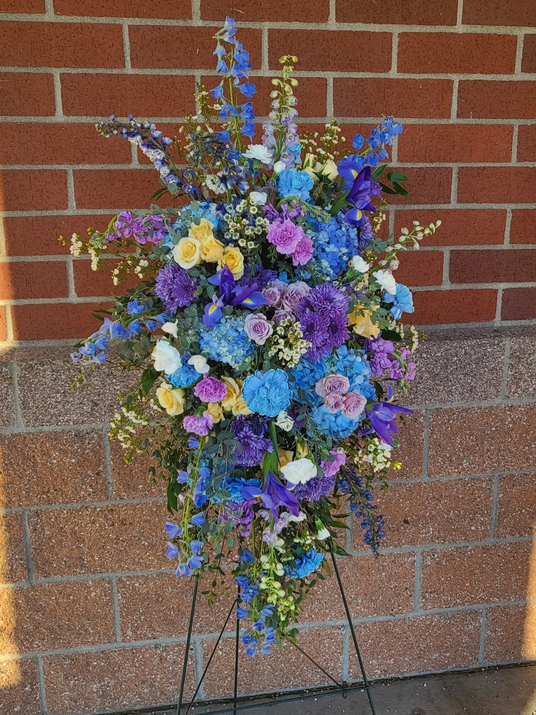 A colorful flower arrangement with blue, purple, yellow, and white flowers is displayed against a red brick wall.
