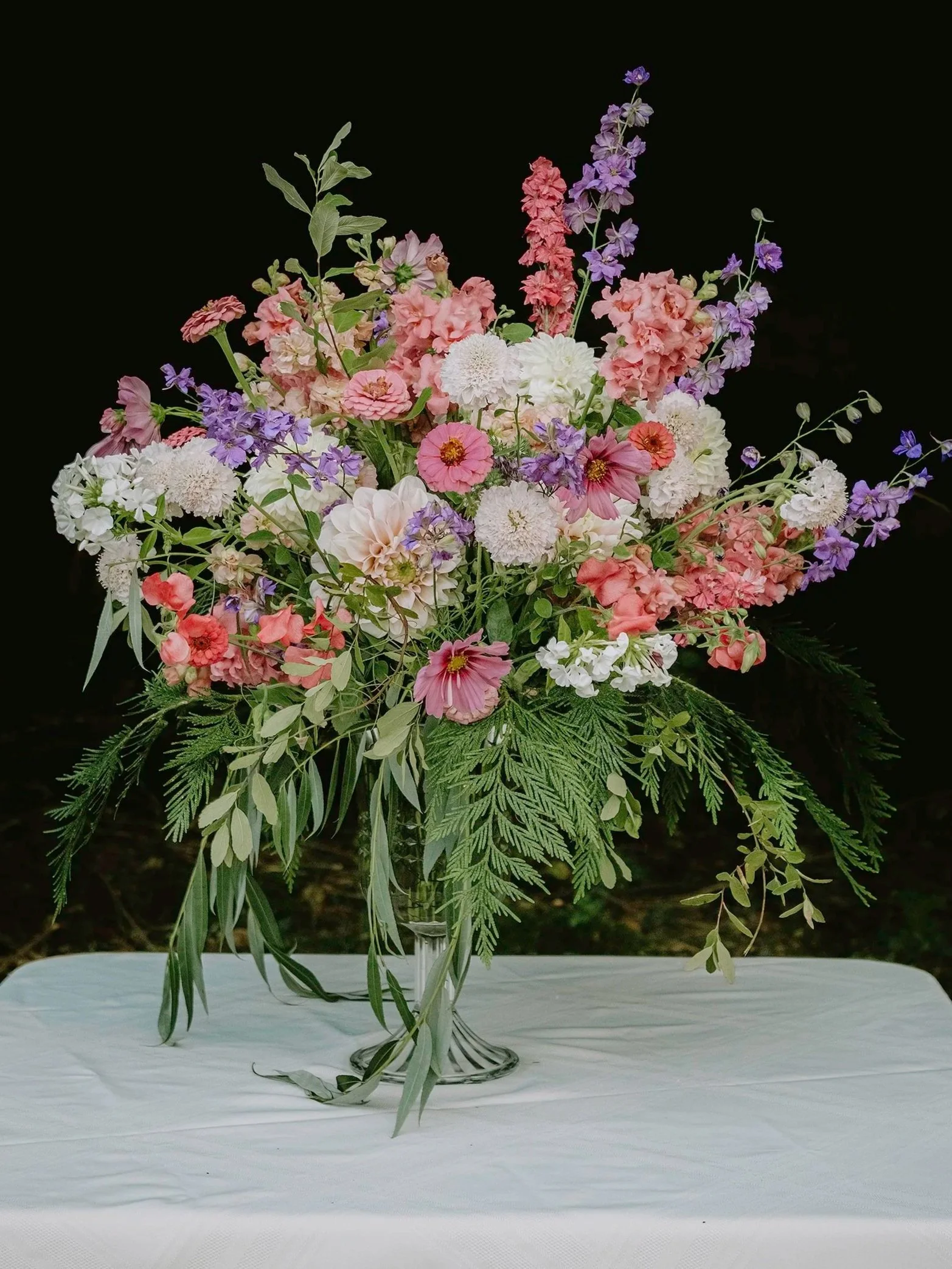 A large floral arrangement in a glass vase on a white table, featuring pink, white, purple, and green flowers and foliage against a black background.