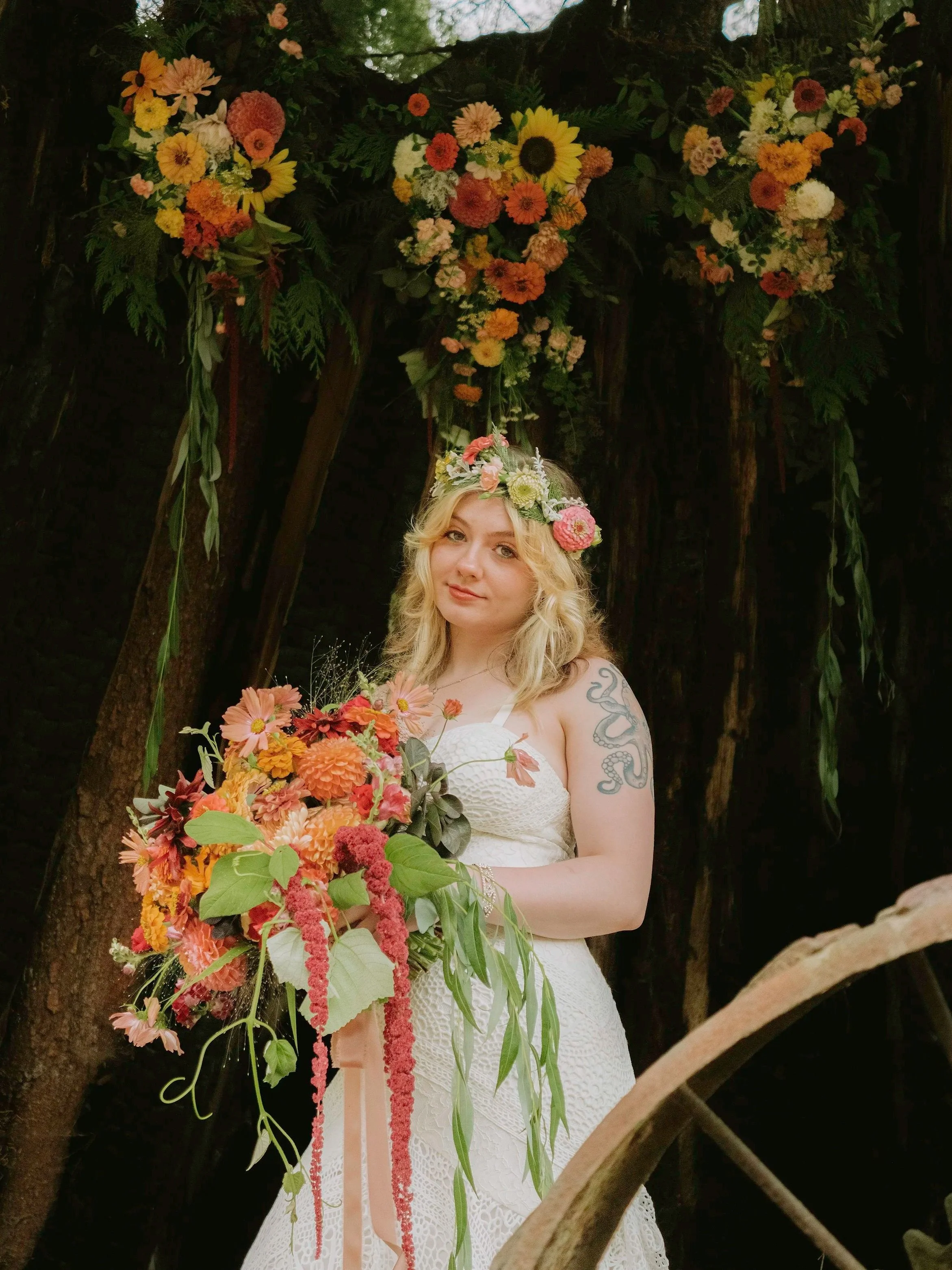 A woman in a white lace dress holding a large bouquet of colorful flowers, standing outdoors beneath a floral arch decorated with various flowers and greenery.