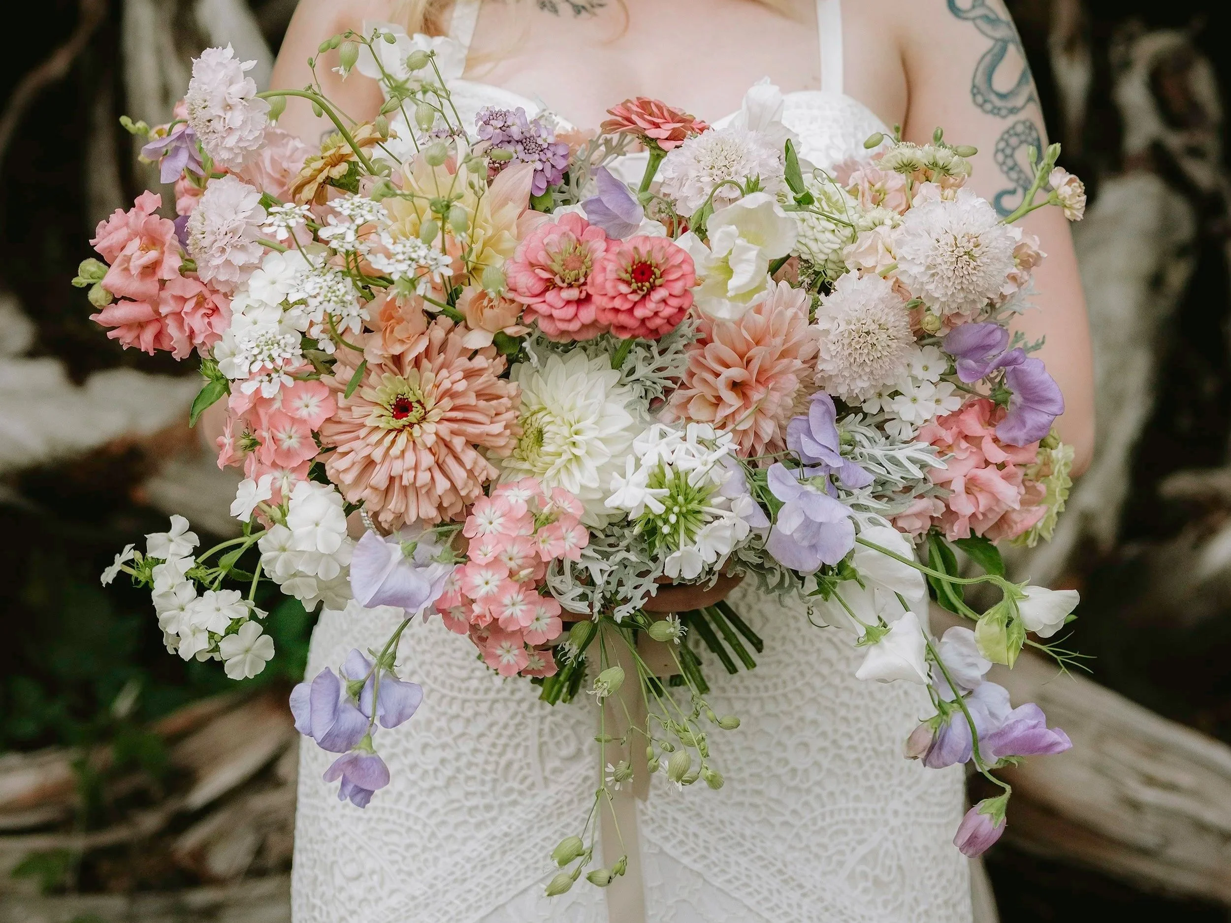 Person holding a large bouquet of mixed pink, white, and purple flowers in front of their torso, wearing a white lace dress, with tattoos on their arms, and a blurred background of branches.