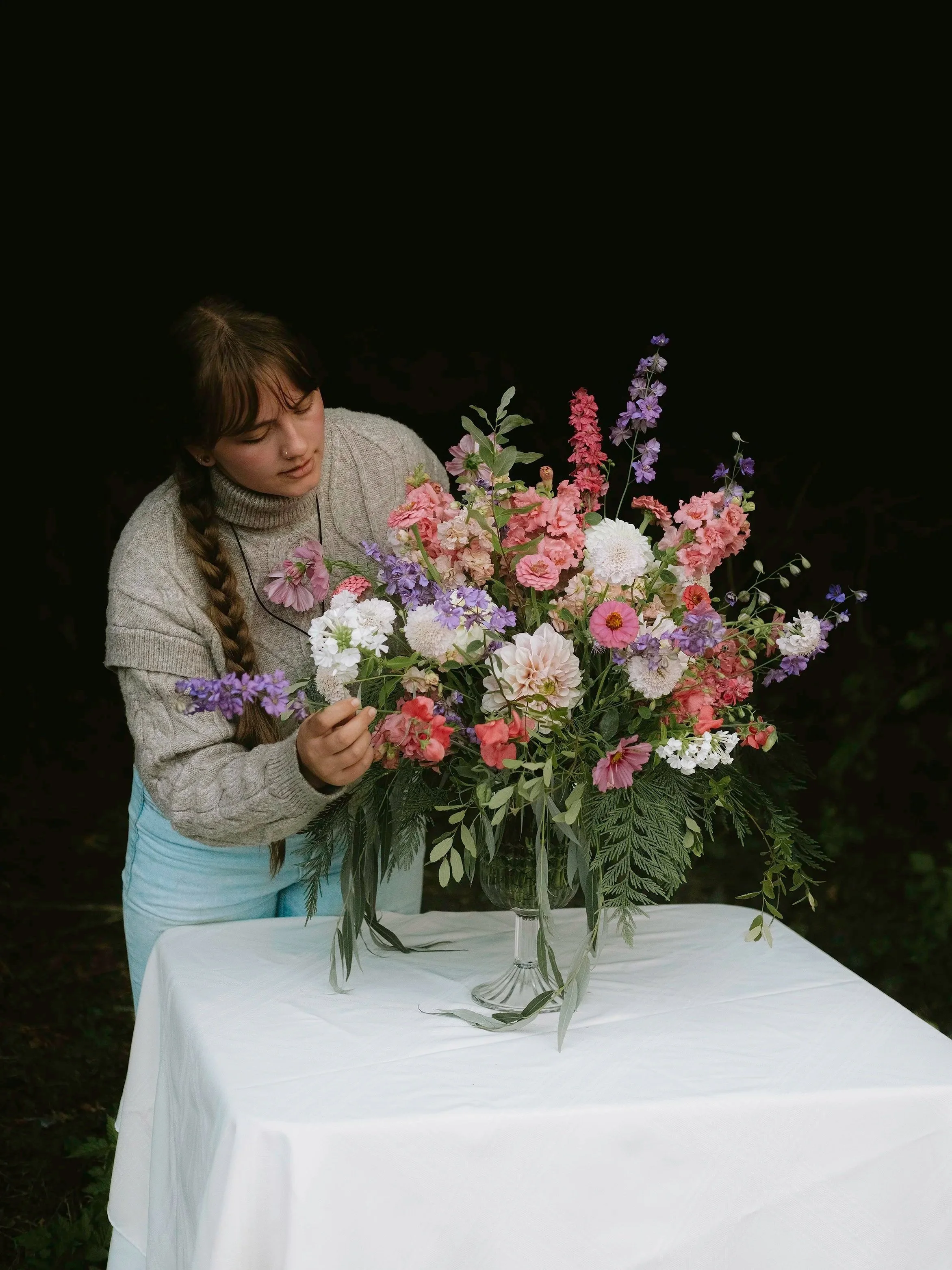 A woman arranging a large, colorful floral bouquet in a glass vase on a white table outdoors.