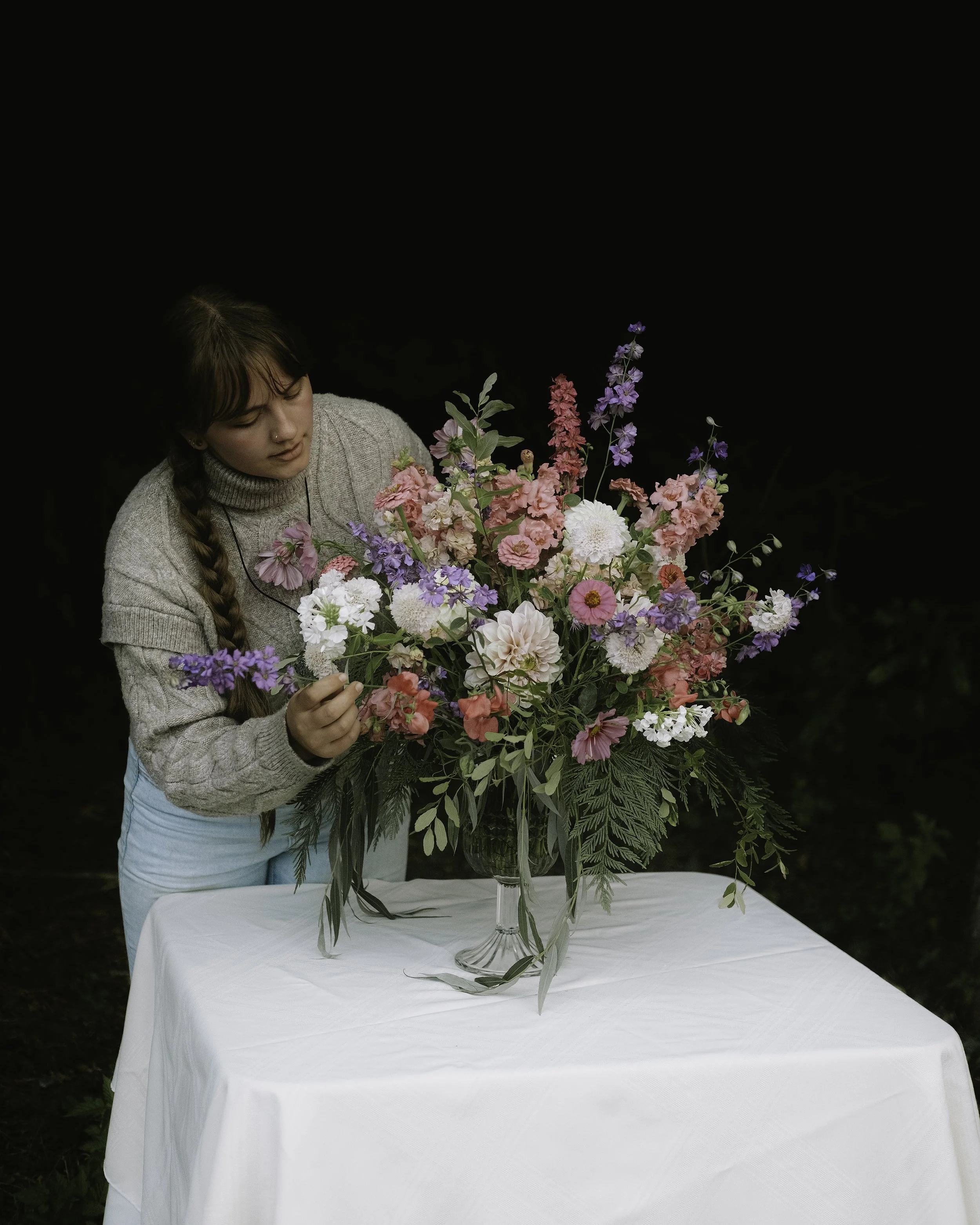 A young woman with long braid in a cozy gray sweater arranging a colorful bouquet of flowers on a white table outdoors against a dark background.