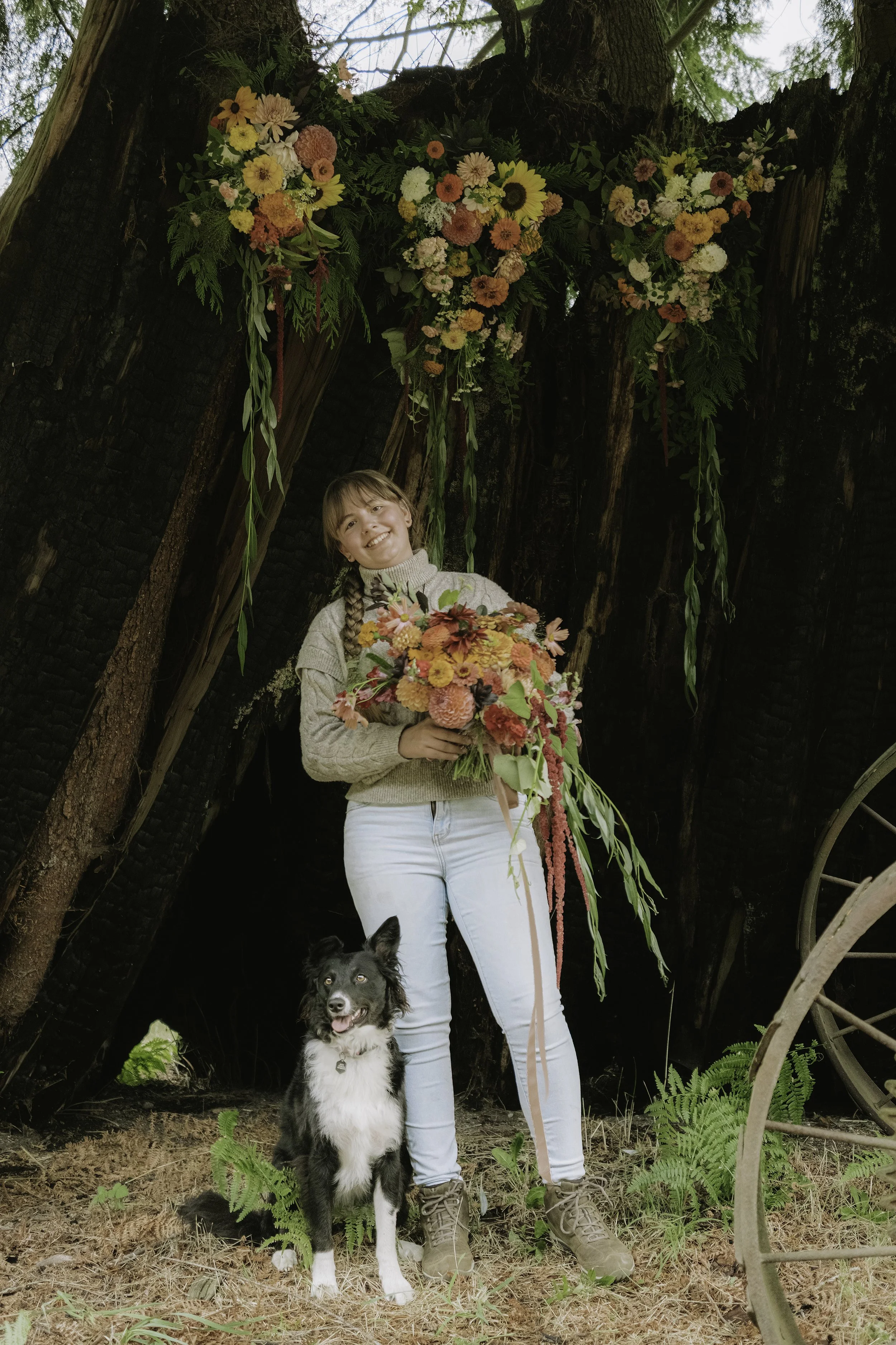 A young woman with braided hair, wearing a beige sweater and white jeans, stands outdoors in front of charred trees, holding a vibrant bouquet of flowers. A black and white dog is sitting on the ground beside her. A large floral arrangement hangs from the trees above.