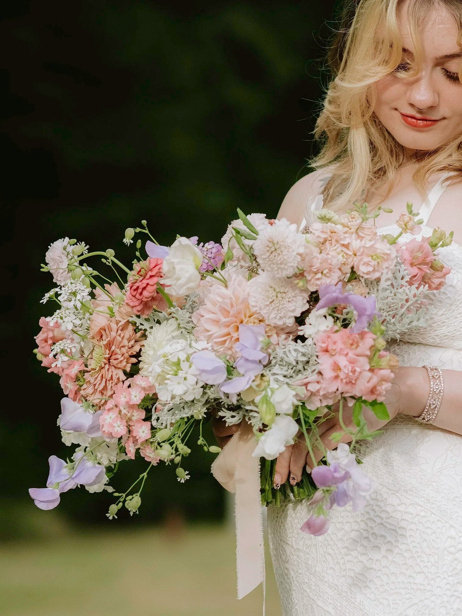 A woman in a white dress holding a large bouquet of pastel-colored flowers, with her eyes closed and a gentle smile.