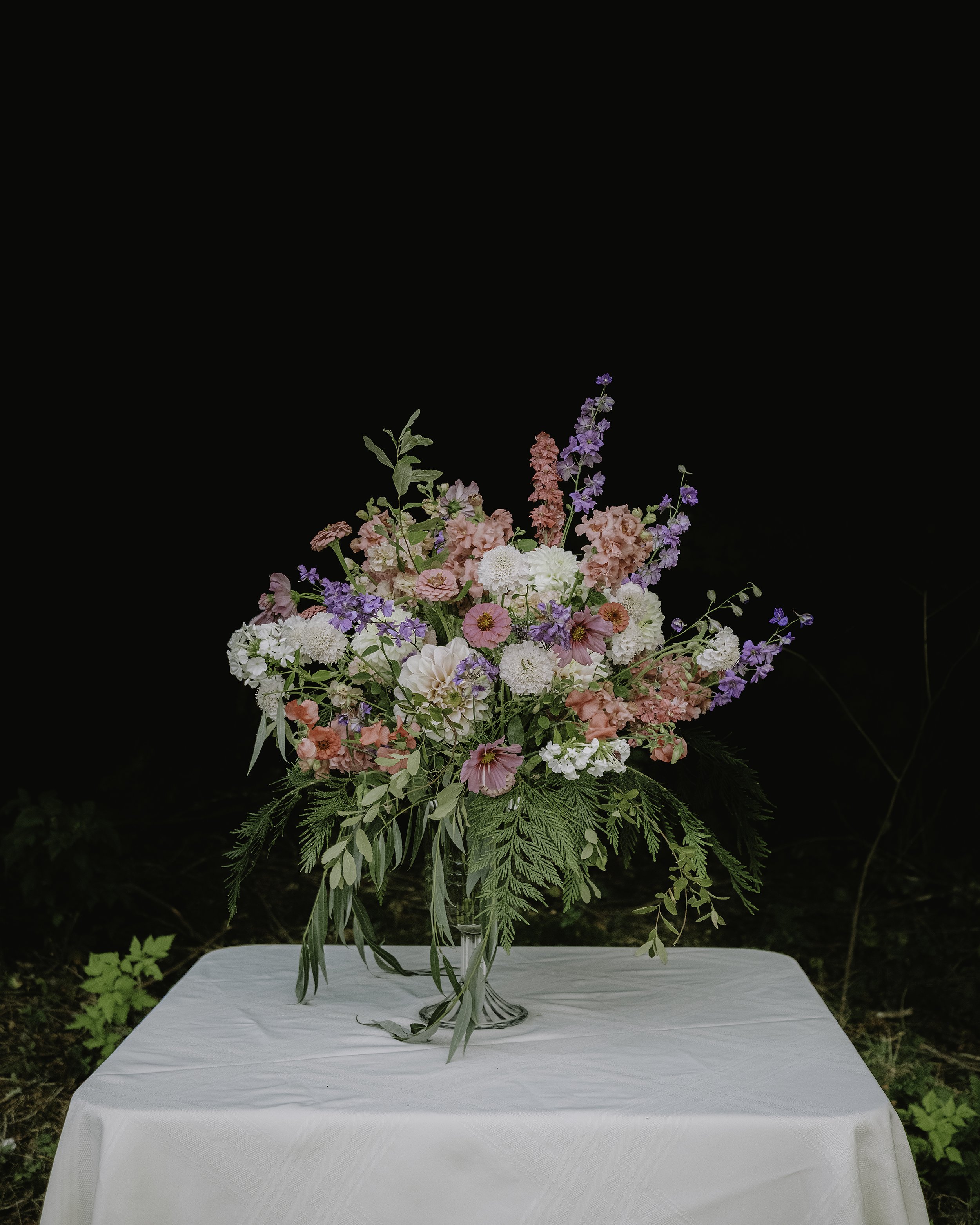 A tall flower arrangement featuring pink, white, and purple flowers with green foliage, placed on a white tablecloth-covered table outdoors with greenery in the background.