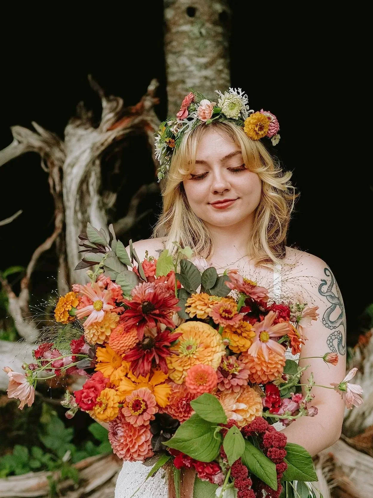 A young woman with blonde hair and a floral crown on her head, holding a large bouquet of orange, pink, and red flowers, with a tattoo on her left arm, standing outdoors against a dark background with a tree trunk.