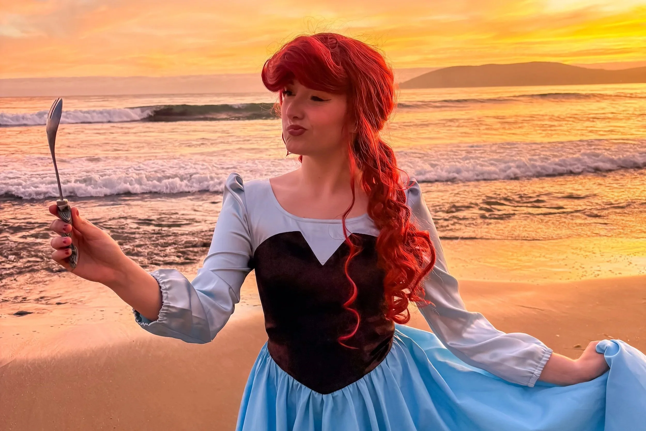 A woman with long red hair is performing a yoga pose on a beach at sunset, holding a spoon in her right hand.