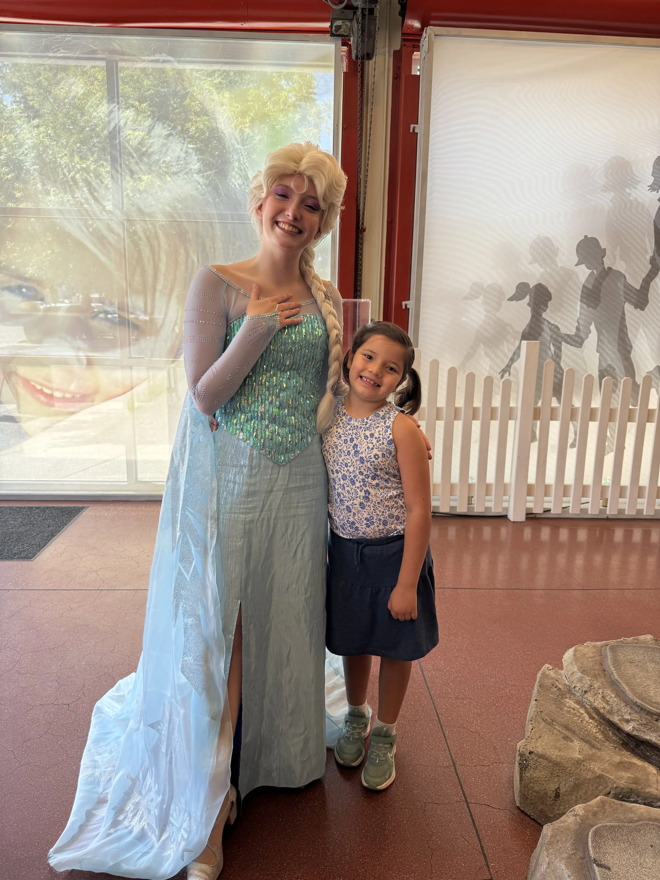 A woman dressed as Elsa from Frozen standing next to a young girl, both smiling at the camera inside a building with a large window and a painted silhouette mural of children playing outside.