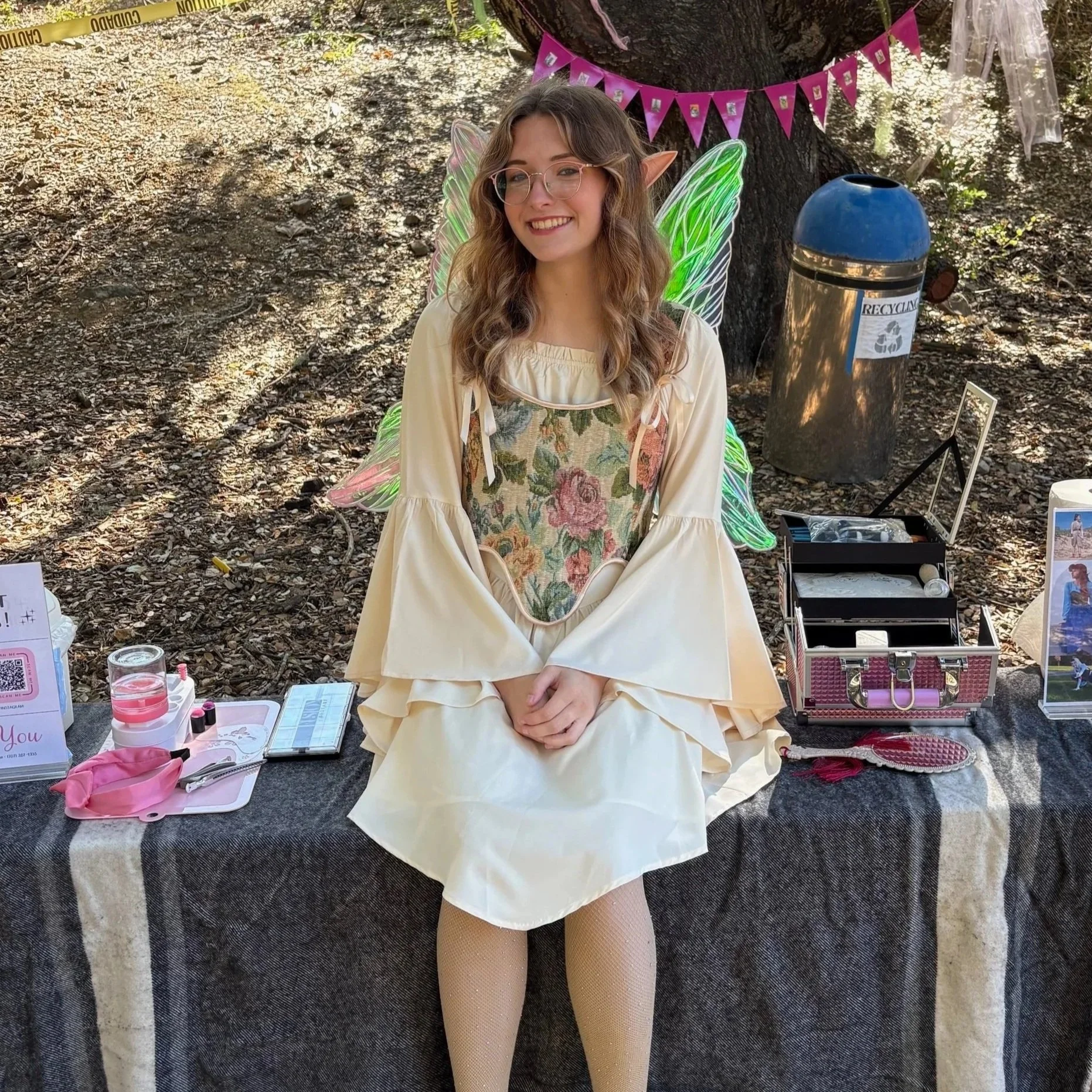 A young woman with elf ears and fairy wings sitting outdoors on a black cloth. She is smiling, wearing glasses, a cream-colored dress, and fishnet tights. There are various makeup items and a pink makeup case on the table in front of her, and a ringed trash can and pink bunting hanging on a tree behind her.
