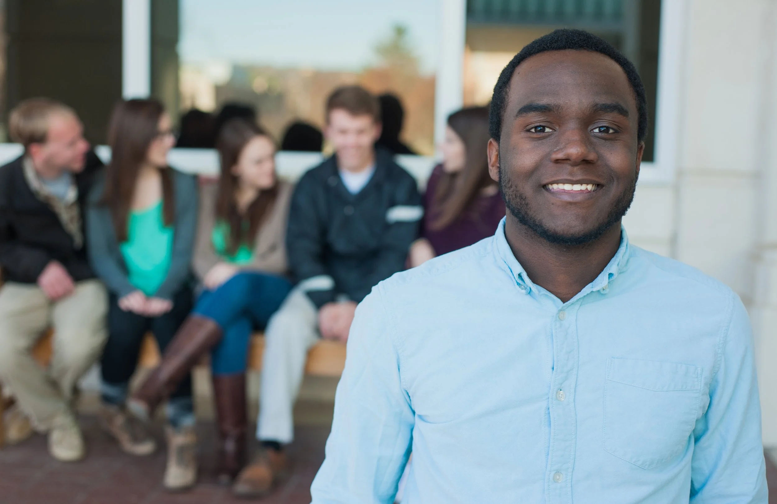 Portrait of a smiling young man wearing a light blue shirt, with a group of people sitting and chatting in the background.
